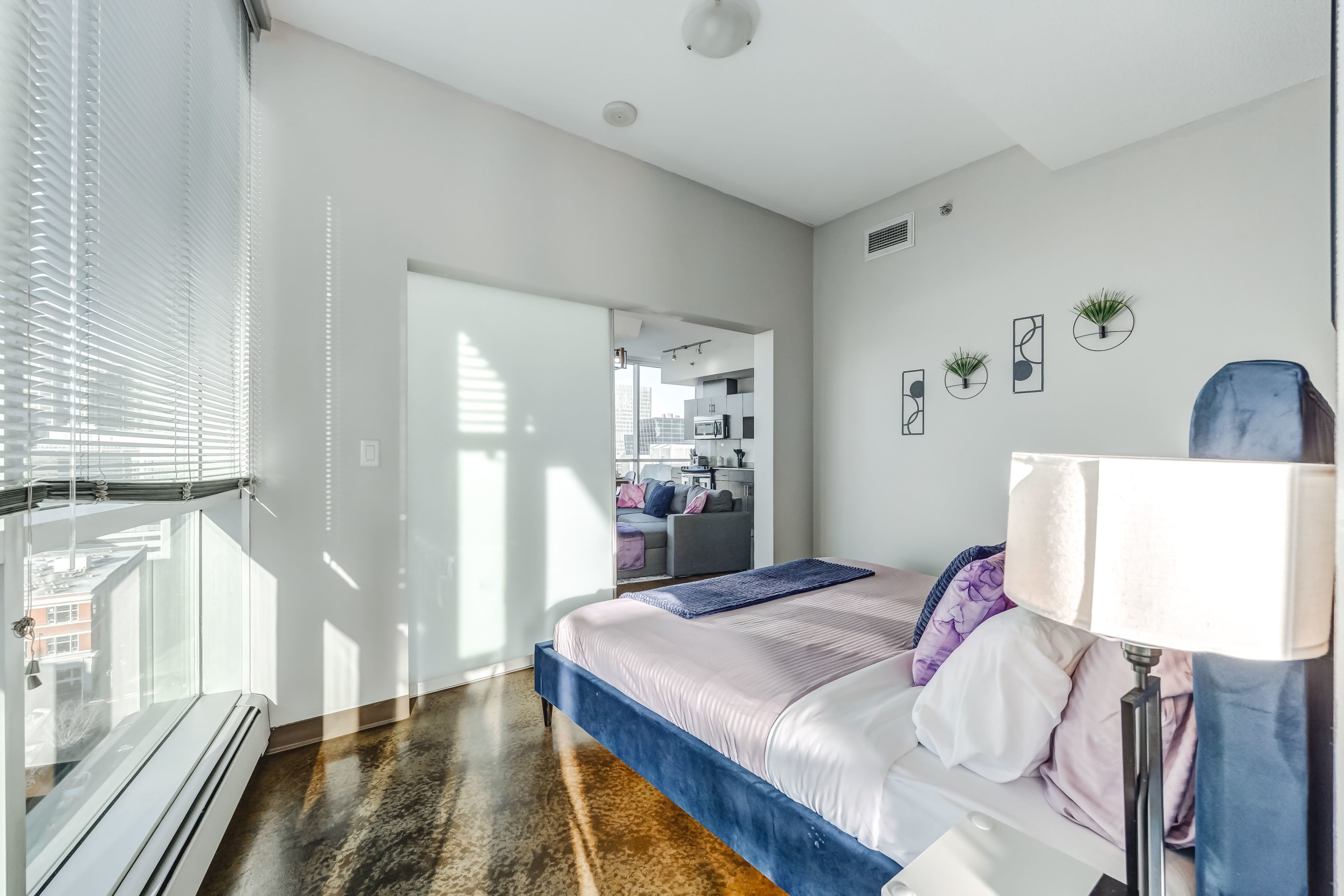 Bright bedroom with a blue velvet bed frame and light pink bedding, featuring a white nightstand with a lamp. A frosted glass sliding door connects to the living area, and large windows with blinds offer natural light.