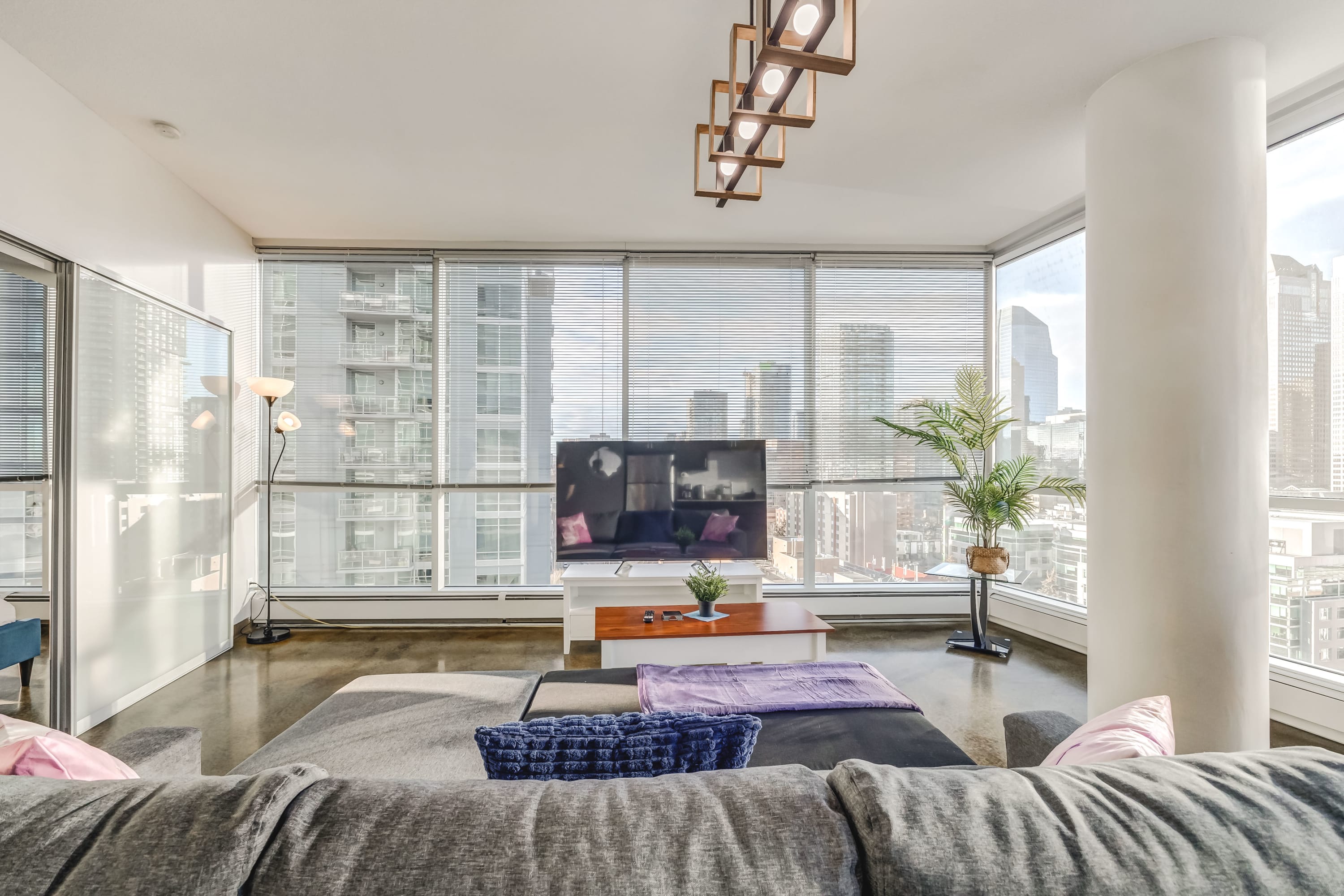 Living room featuring a gray sectional sofa facing a large flat-screen TV, with expansive floor-to-ceiling windows covered by blinds. A dining area is visible to the right, and a bedroom is accessible via a sliding door on the left.