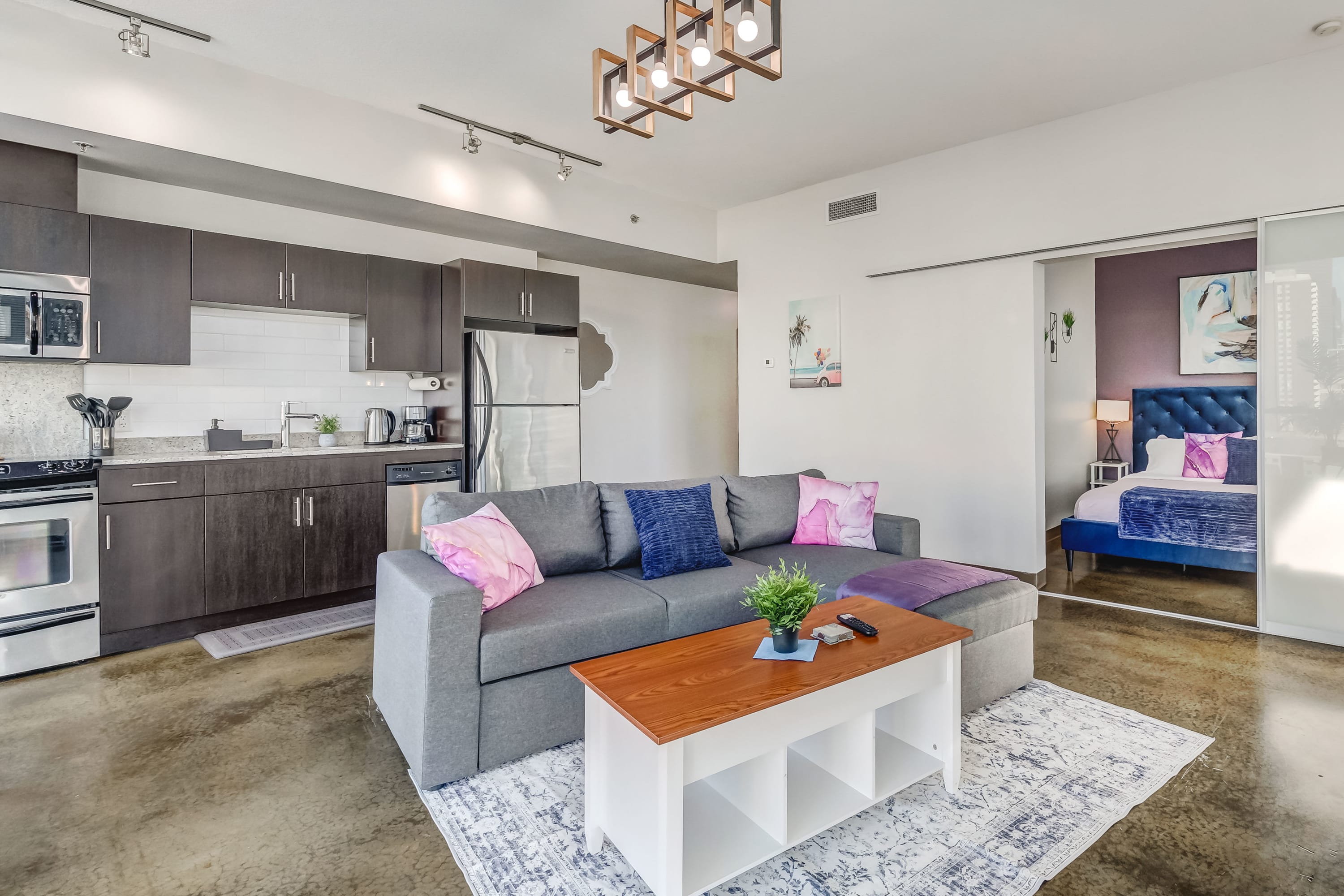 View of the open-concept living room featuring a gray sectional sofa, white and wood coffee table, and a modern chandelier. The kitchen is visible in the background, and a bedroom entrance is to the right, all on polished concrete floors.