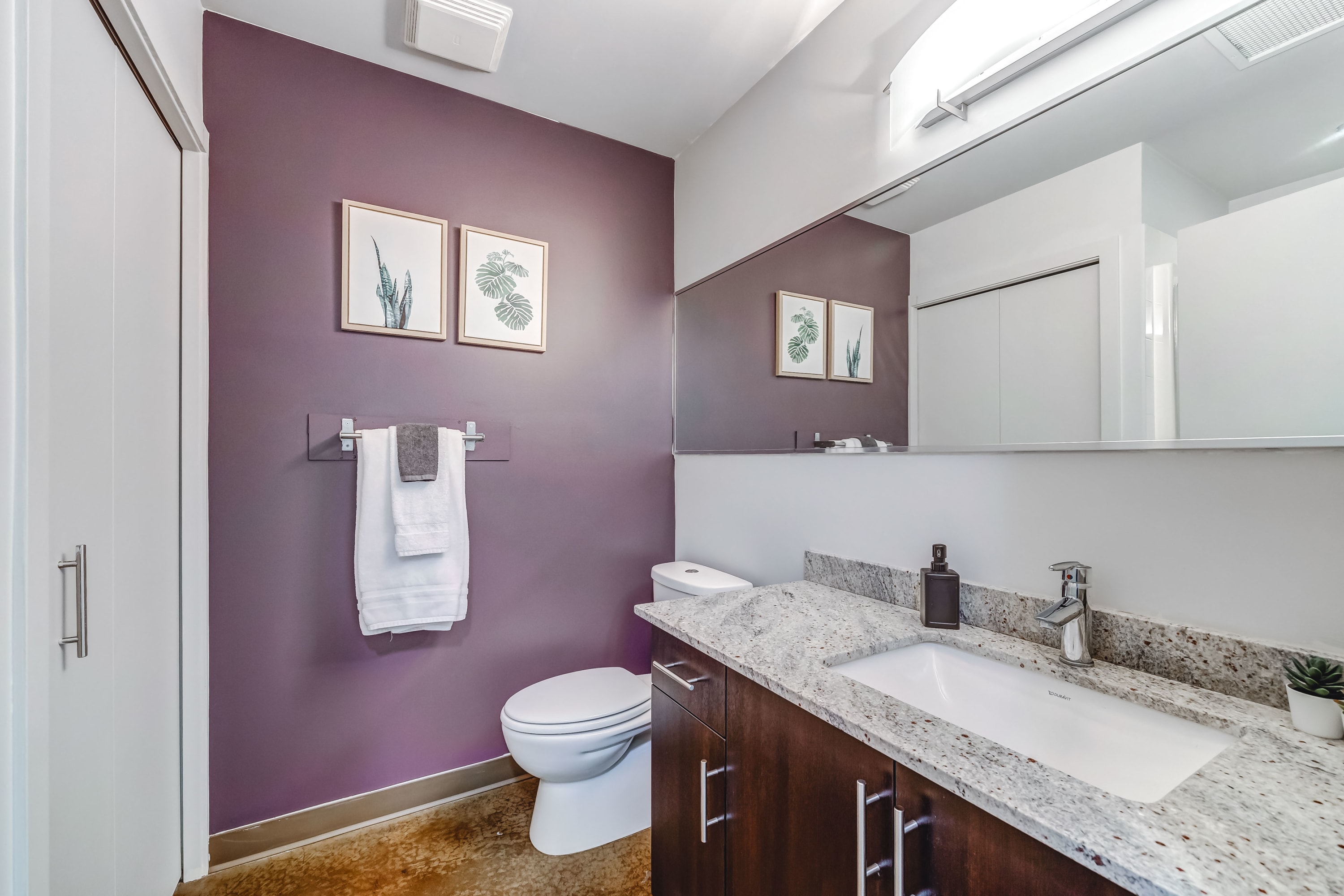 Modern bathroom with a dark purple accent wall, a white toilet, and a vanity with a granite countertop and an undermount sink. A large mirror extends across the wall, reflecting the room.