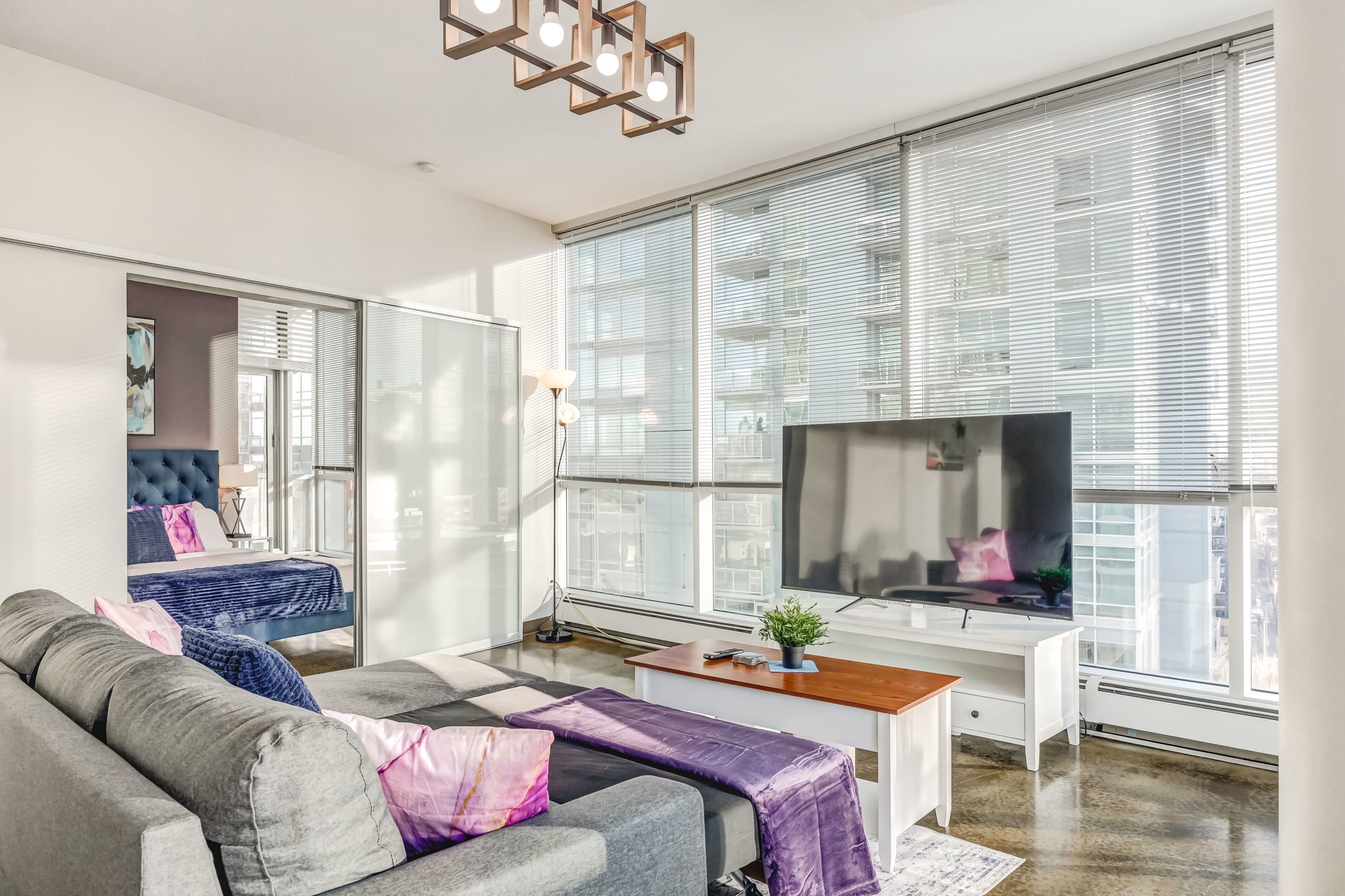 Modern living room with a grey sectional sofa, a white TV stand with a large flat-screen TV, and floor-to-ceiling windows with blinds offering city views. A glimpse of a bedroom with a blue bed is visible through a sliding frosted glass door.