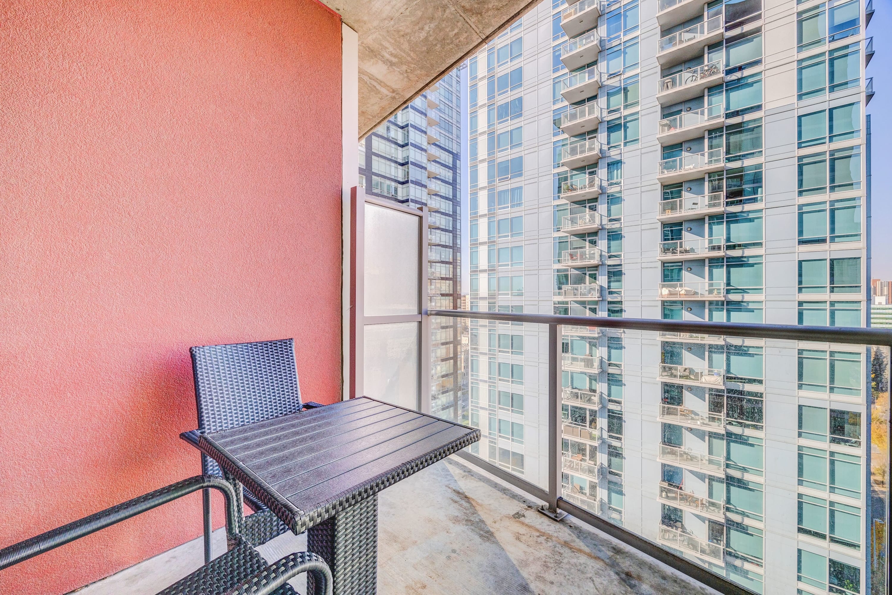 Private balcony with a small black wicker table and chairs, set against a vibrant red accent wall. It offers views of adjacent modern high-rise buildings through a glass railing.
