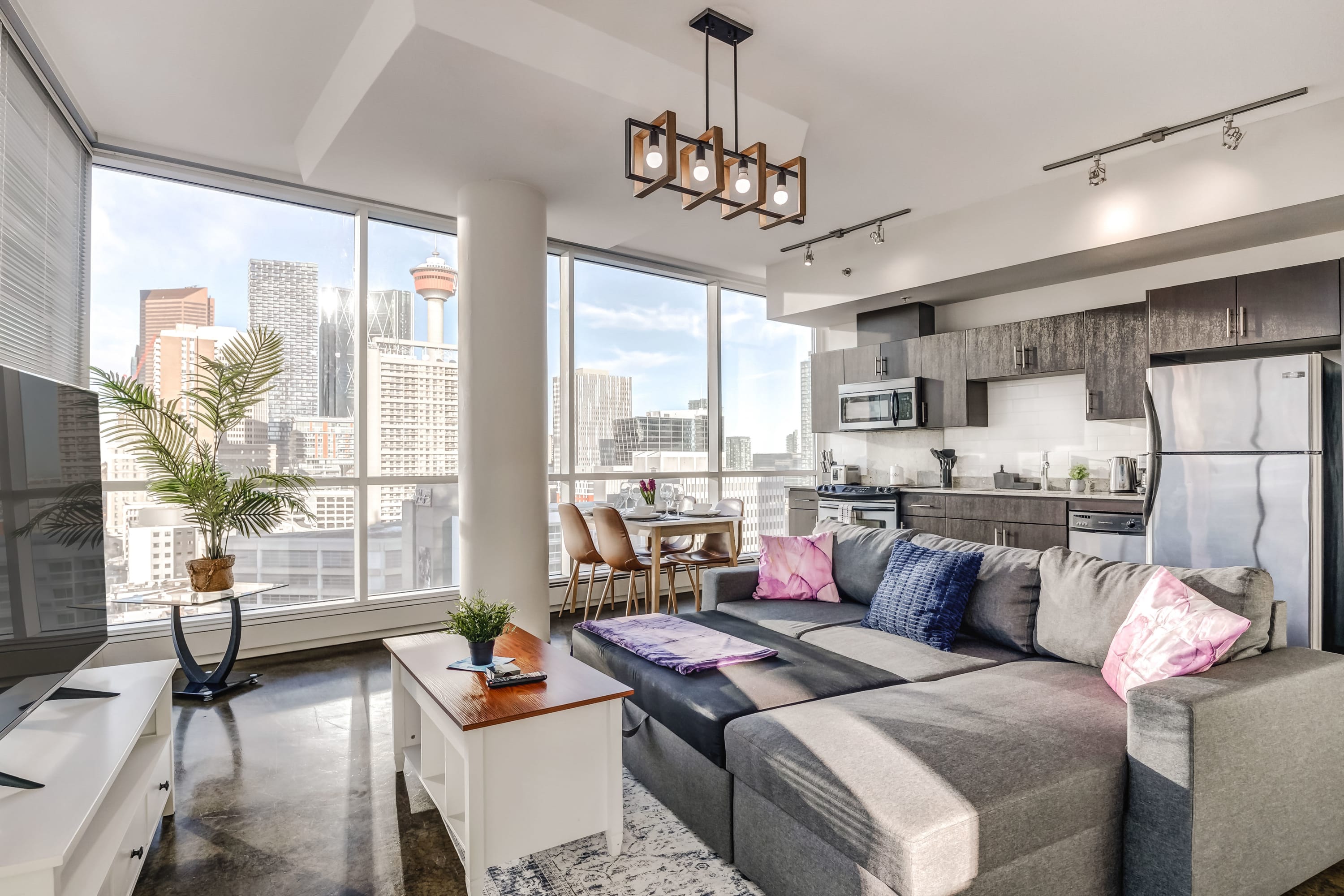 Bright open-concept living area with a gray sectional sofa, white coffee table, and a modern chandelier. Floor-to-ceiling windows showcase stunning downtown city views, including a prominent tower, over polished concrete floors.