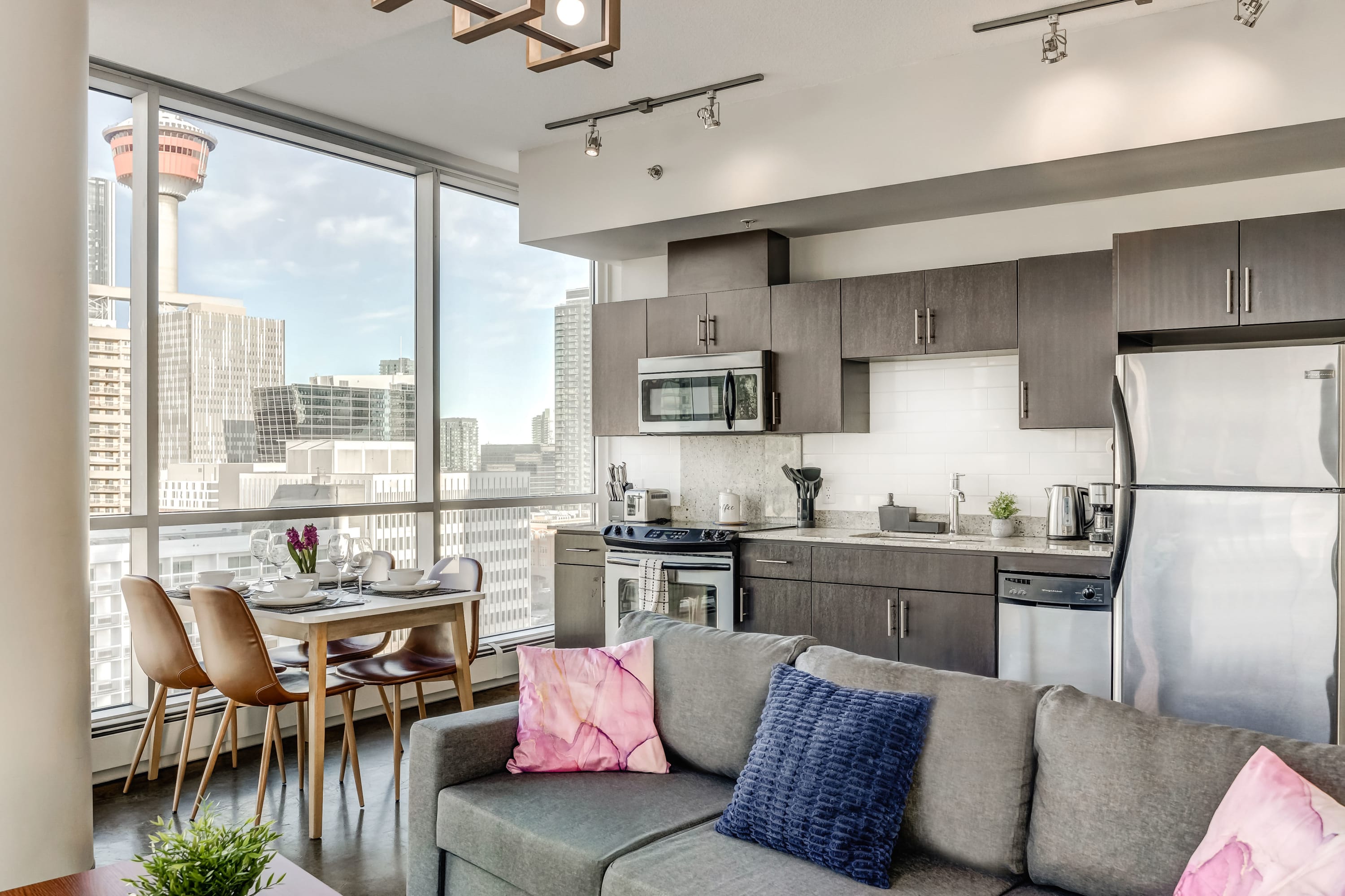 Open-concept living space featuring a grey sectional sofa, a dining area with a wooden table and four chairs, and a modern kitchen with dark wood cabinets and stainless steel appliances. Large windows offer panoramic city views, including a prominent tower.