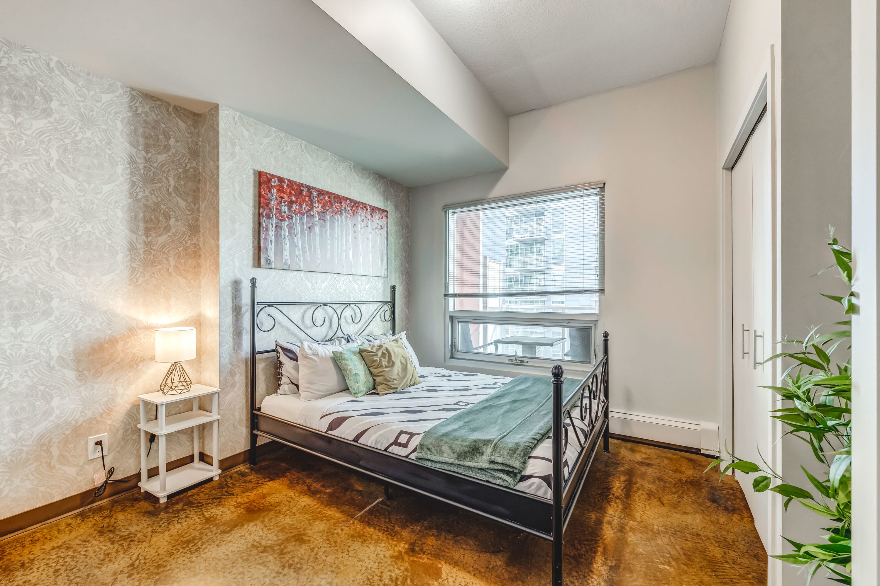 Second bedroom with a black wrought iron bed frame, patterned bedding, and a white nightstand with a lamp. The room has a textured accent wall and a window with blinds, providing natural light.