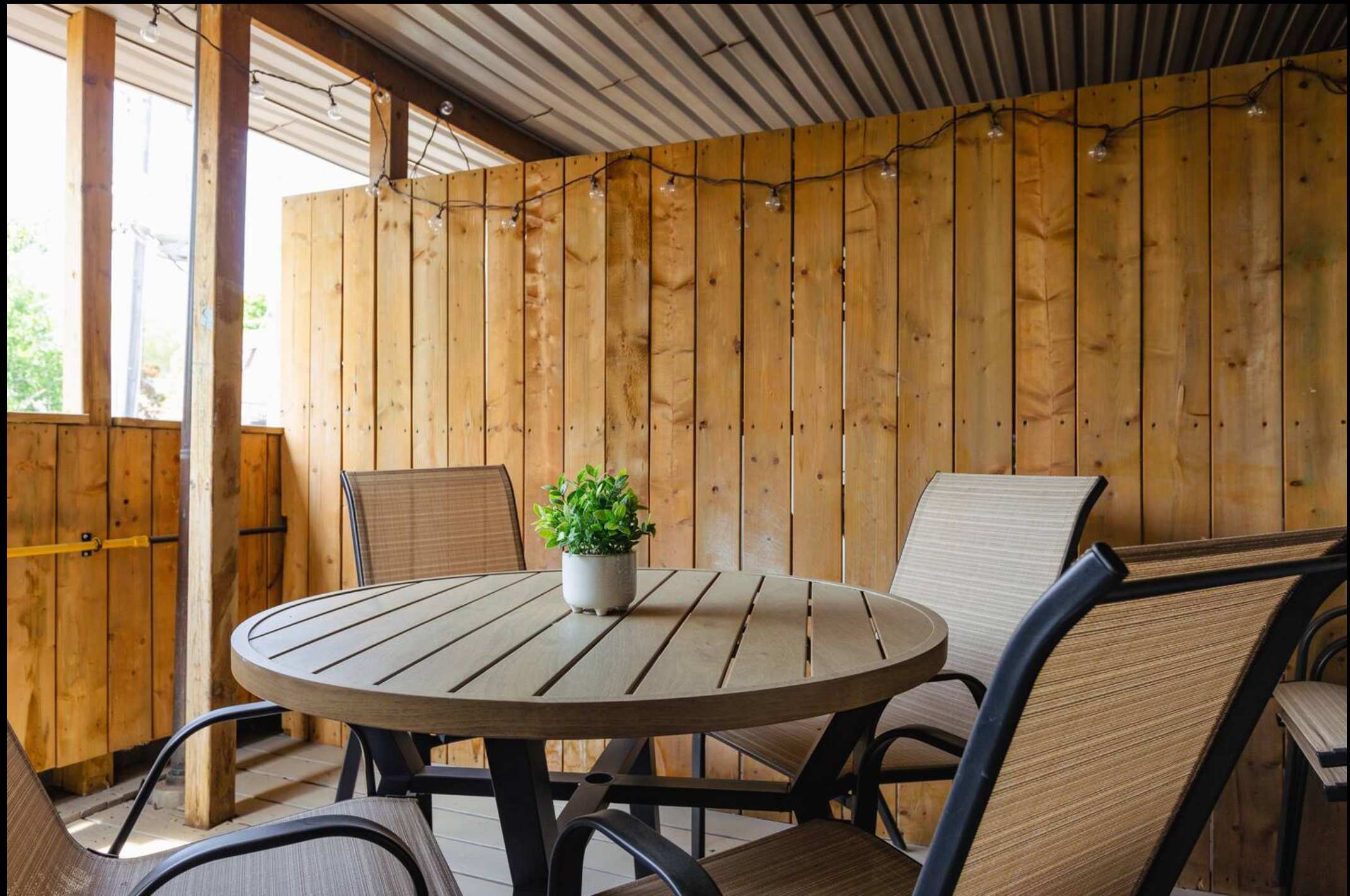 Close-up view of the covered outdoor patio, highlighting a round wooden dining table with a potted plant centerpiece and four mesh-backed chairs, surrounded by wooden slatted walls with string lights.