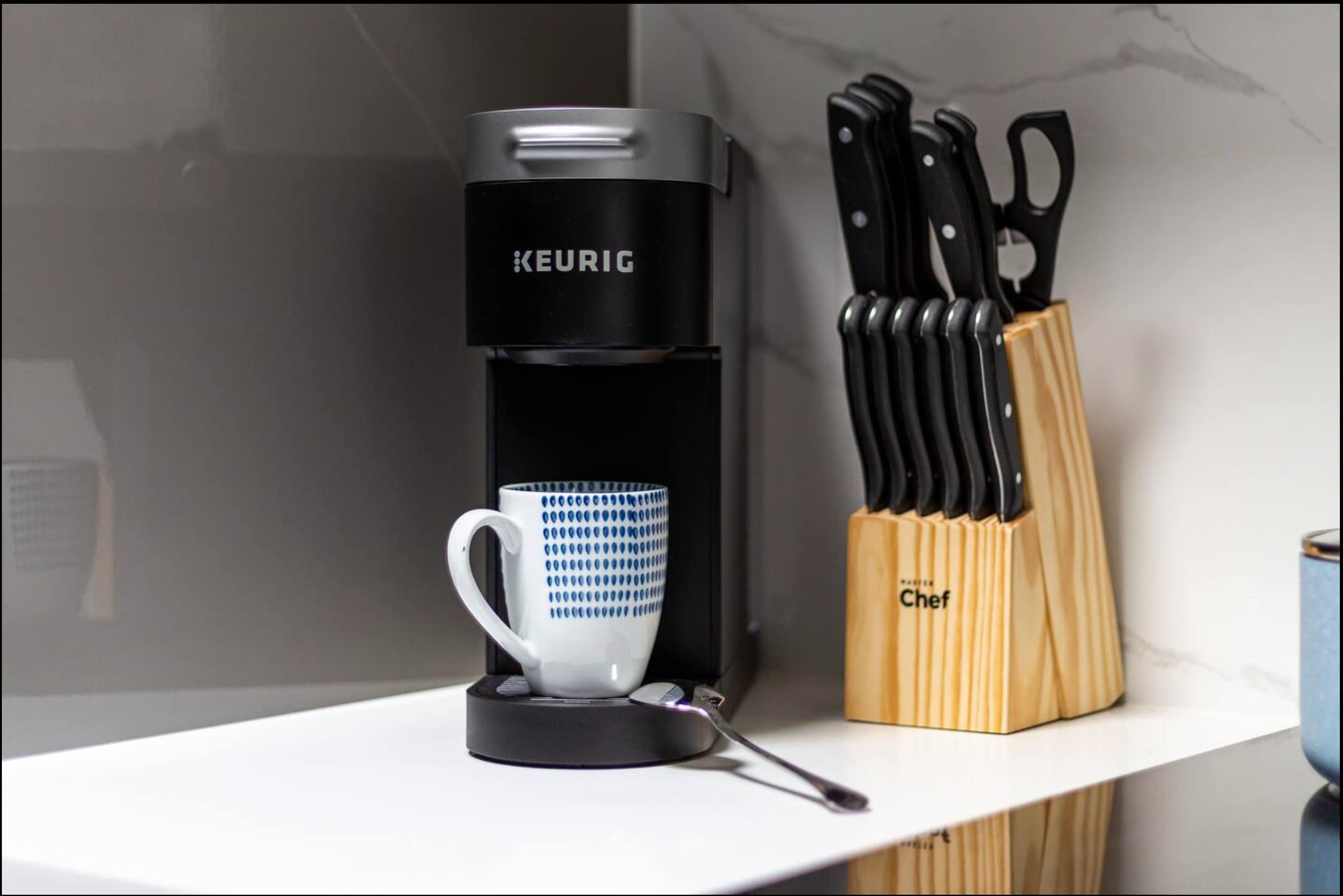 Close-up view of a black Keurig coffee maker with a blue and white patterned mug, positioned next to a wooden knife block on a white countertop with a marble-patterned backsplash.