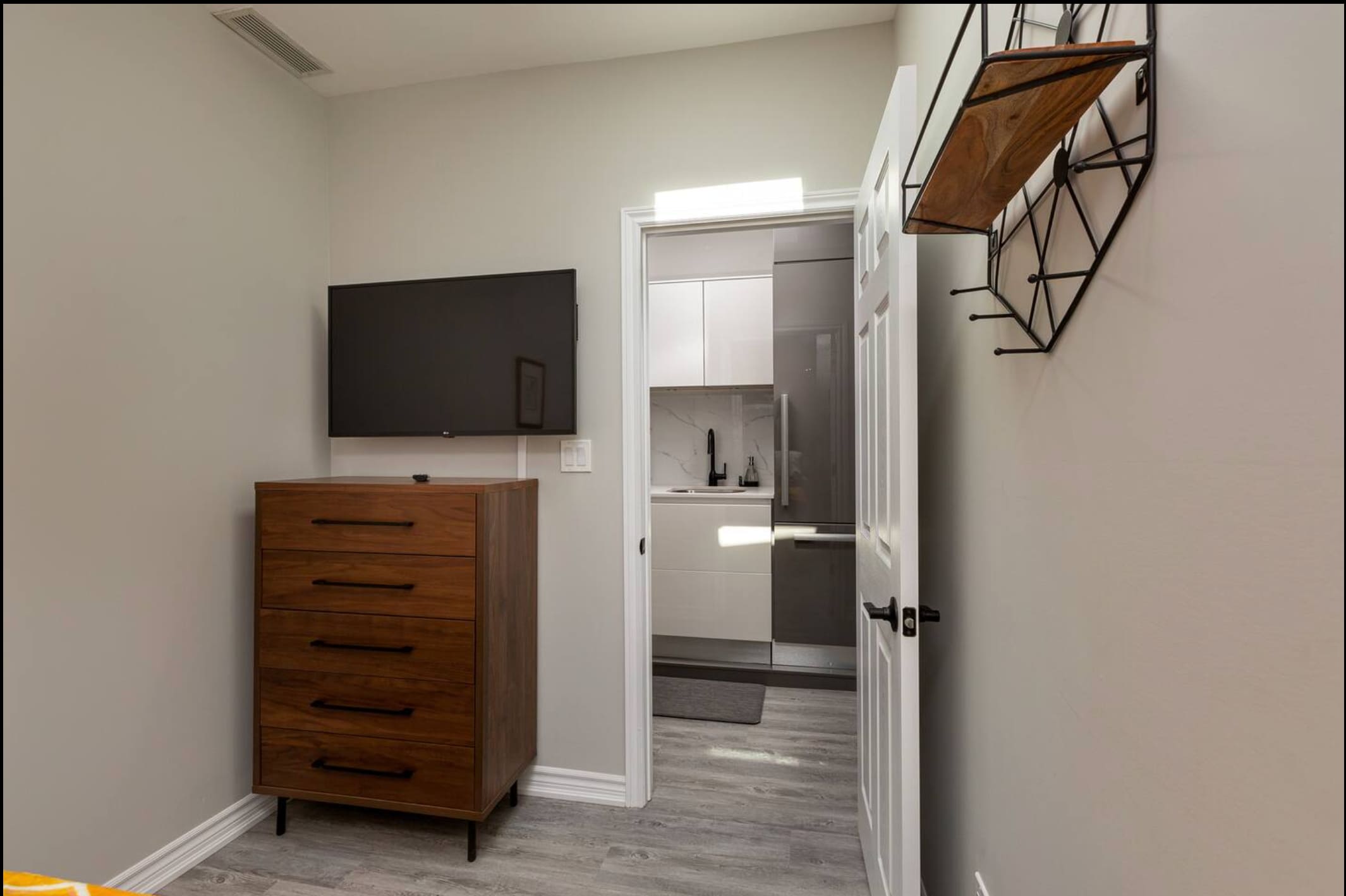 View of the bedroom featuring a wall-mounted flat-screen TV above a modern wooden dresser with black handles. An open doorway on the right reveals a glimpse of a compact kitchen with white and gray cabinetry and a refrigerator.