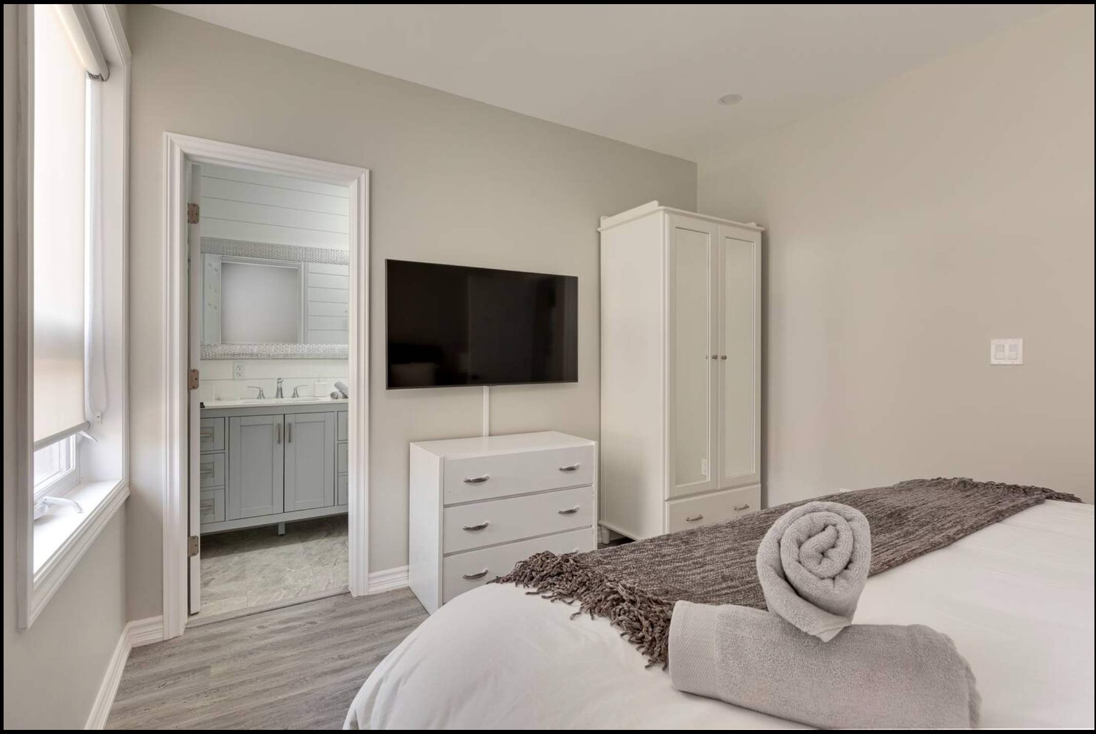 Bedroom view showing a wall-mounted flat-screen TV above a white dresser, a white wardrobe, and an open doorway leading into a modern bathroom with a vanity and mirror.