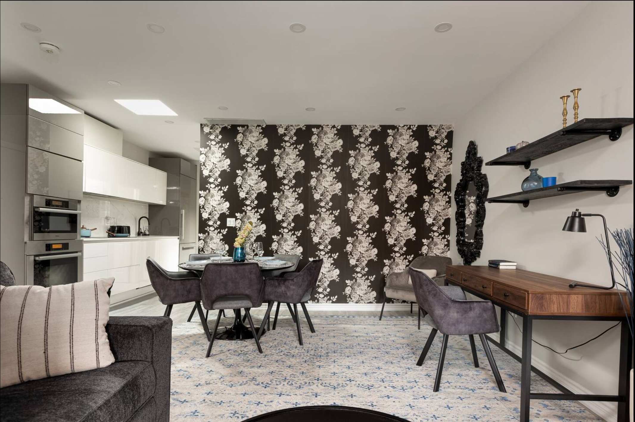 Open-concept living and dining area with a striking black and white floral accent wall, a round dining table with gray velvet chairs, and a dedicated workspace with a wooden desk and chair.