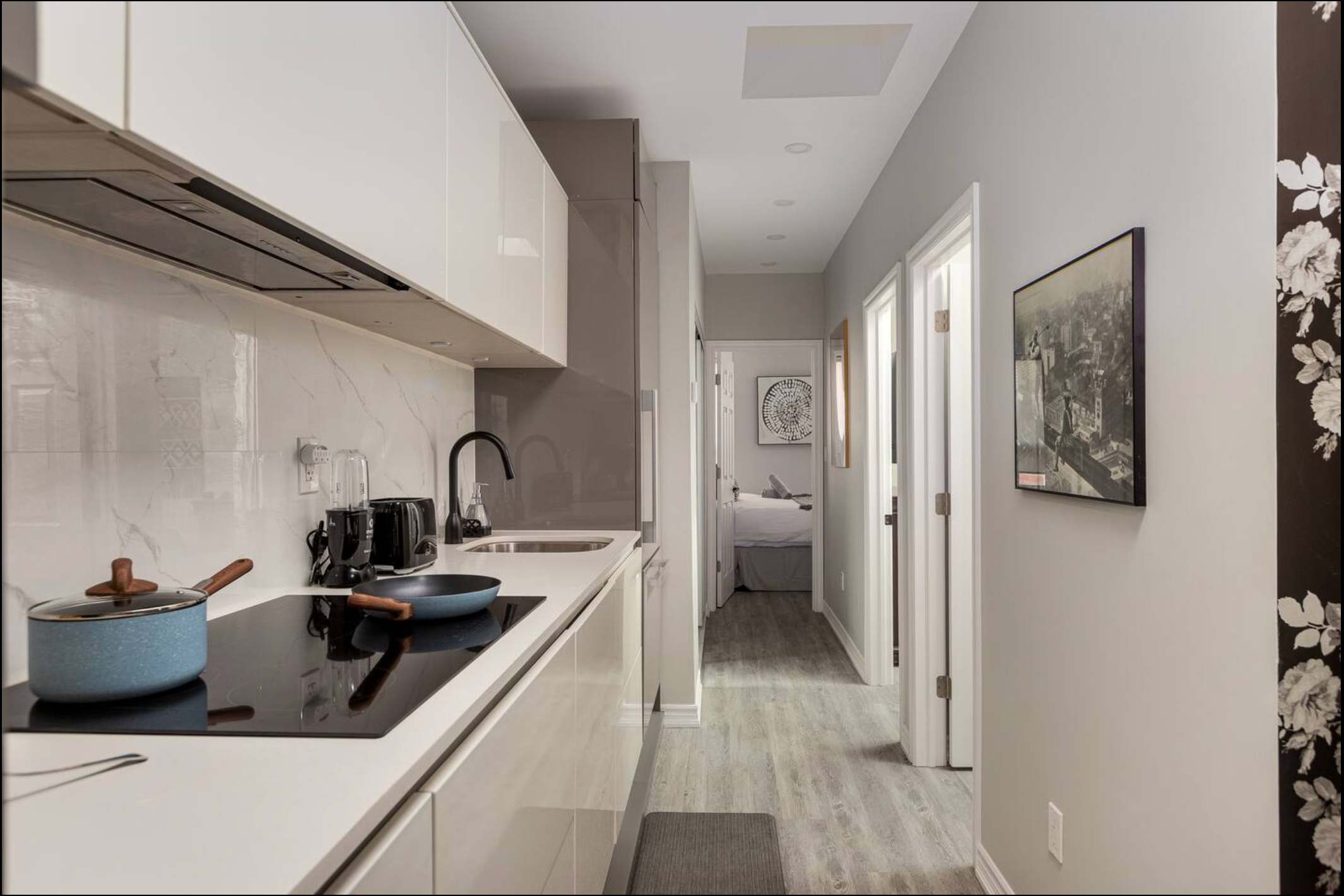 View of the modern kitchen with white cabinets and marble backsplash, extending into a light gray hallway with light wood-look flooring, leading to a bedroom and bathroom.