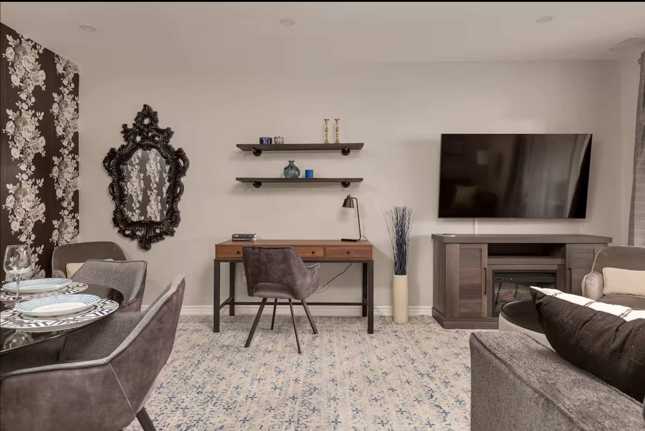 Spacious open-concept area featuring a living room with a TV and media console, an office nook with a wooden desk and chair, and a dining area with a floral accent wall and round table.