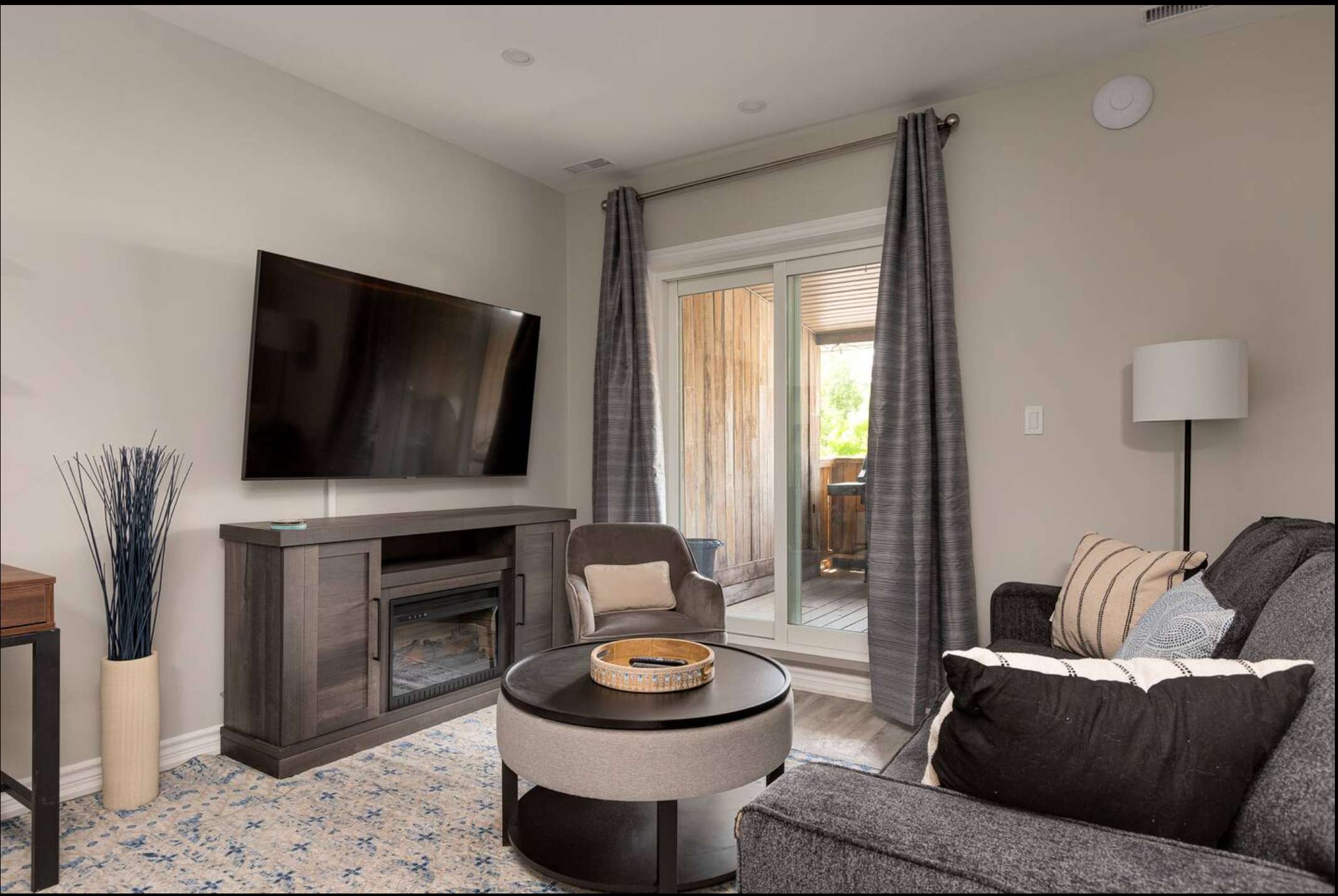 View of the living room from another angle, showing a dark gray sectional sofa with decorative pillows, a media console with a fireplace, and a sliding door to the patio, with an armchair and coffee table.