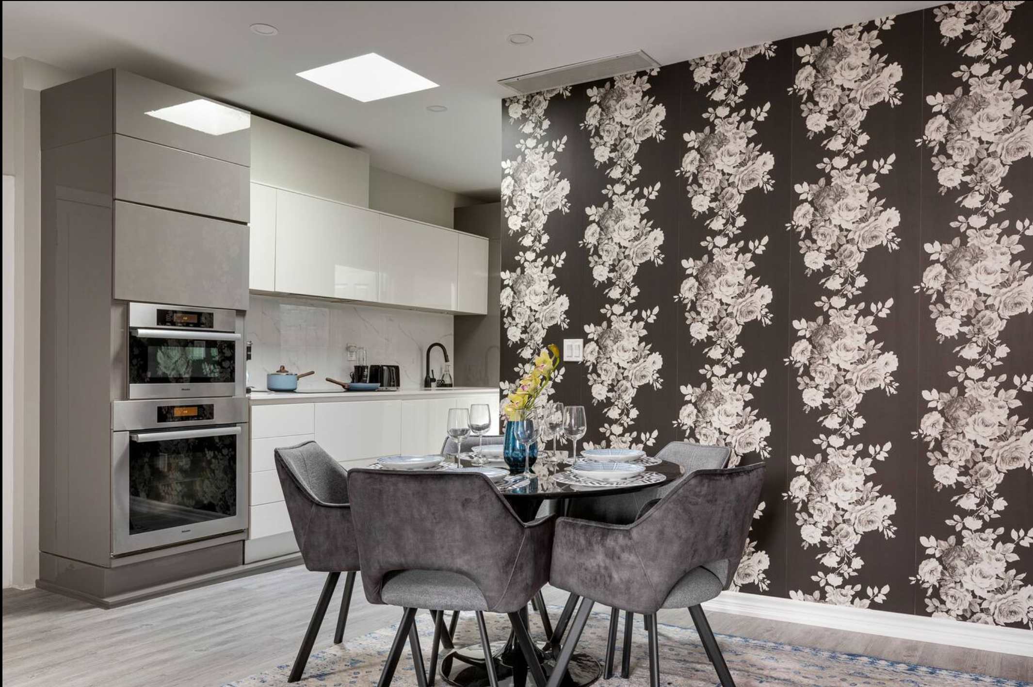 Modern dining area with a black round table and gray velvet chairs, set against a dramatic black and white floral accent wall, with a contemporary kitchen visible in the background.