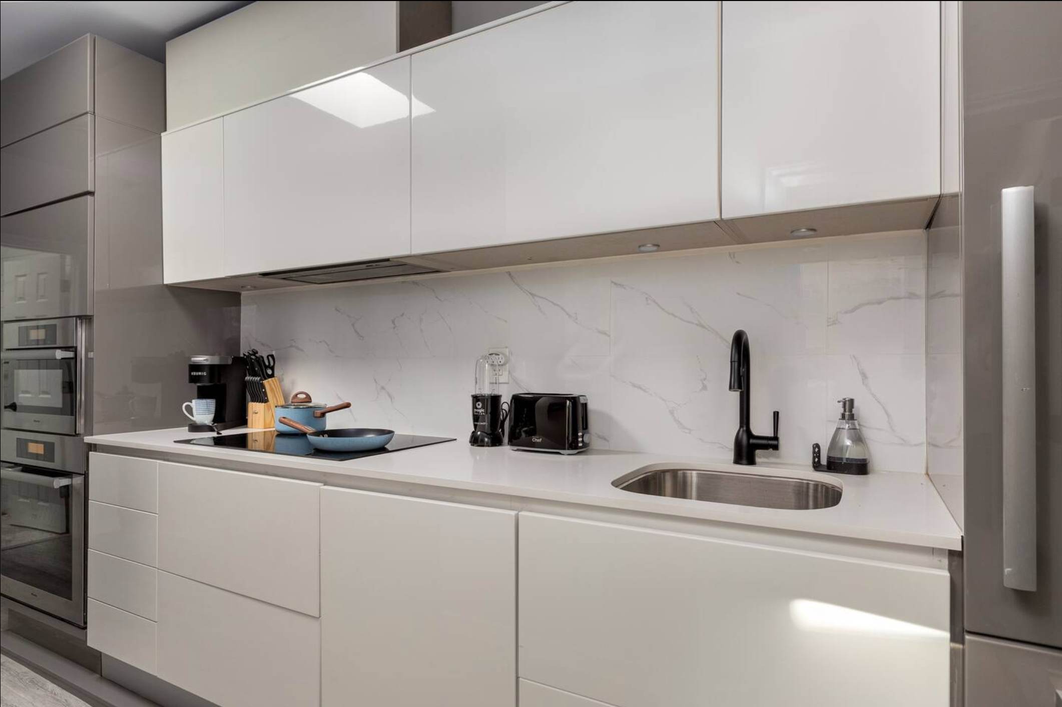 Sleek, modern kitchen with glossy white upper and lower cabinets, a white quartz countertop, and a white marble-patterned backsplash, featuring a black sink, faucet, and induction cooktop.