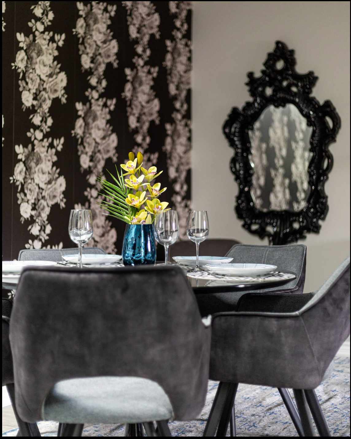 Elegant dining area featuring a round table set with dark gray upholstered chairs, a vibrant floral wallpaper accent wall, and a decorative black ornate mirror reflecting the room.