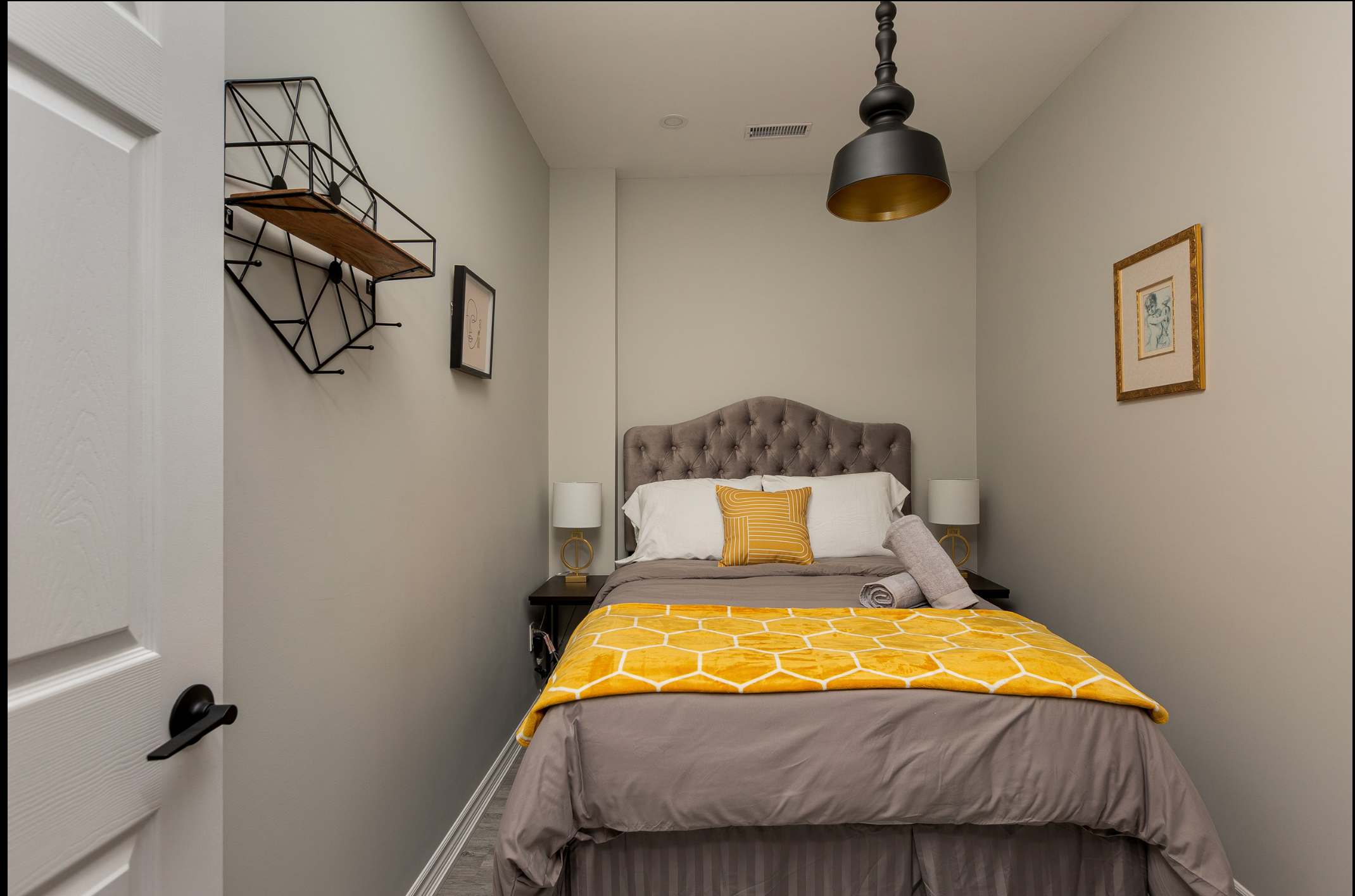 A cozy bedroom with light gray walls, a queen-sized bed featuring a tufted gray headboard, gray bedding, and a vibrant yellow honeycomb-patterned throw blanket. A black geometric wall shelf with a wooden plank is mounted on the left, complemented by a black pendant light.