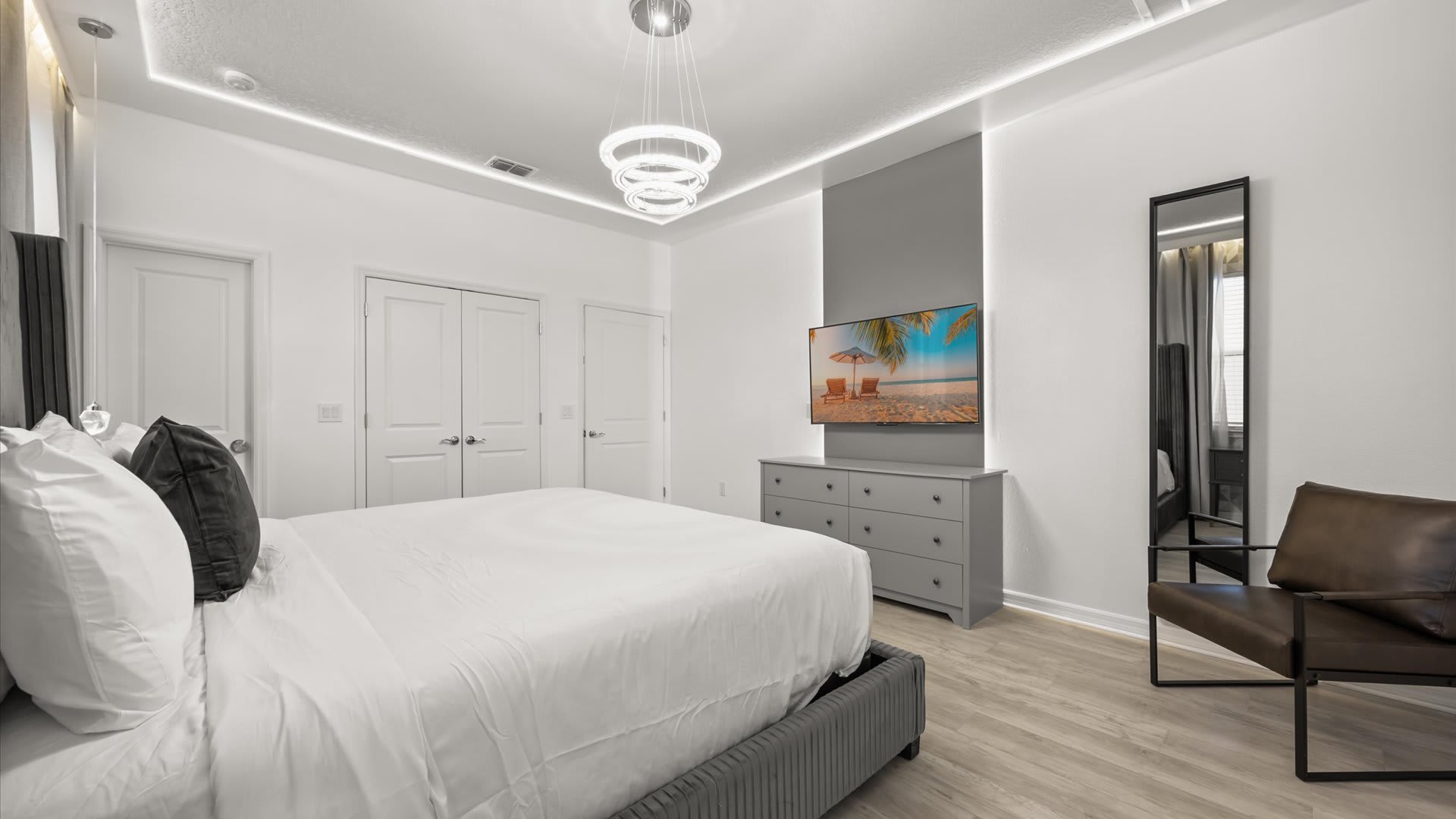 Another view of the modern master bedroom, showcasing a large gray dresser with a wall-mounted TV, a dark leather accent chair, and a full-length mirror, with light wood-look flooring throughout.
