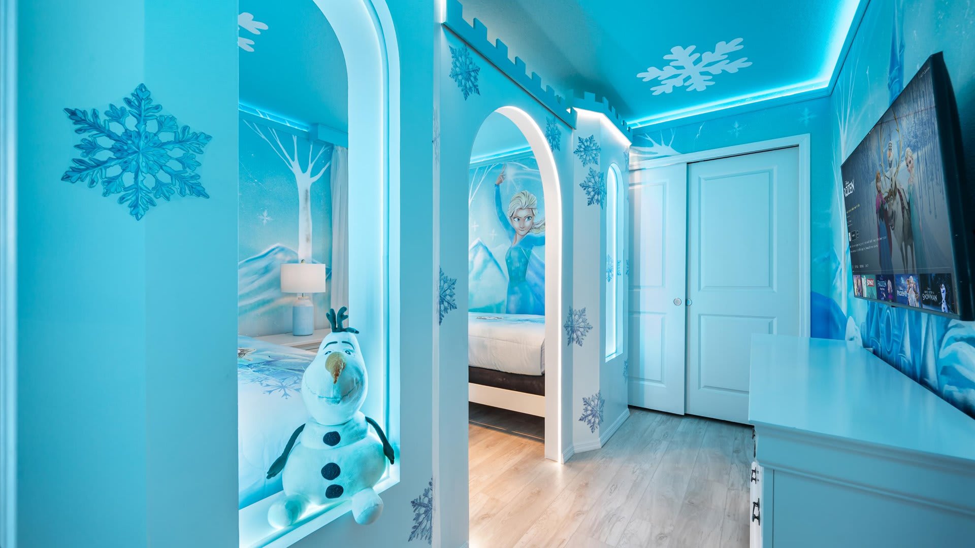Enchanting "Frozen" themed bedroom with blue walls, snowflake decorations, and a central archway leading to another sleeping area, featuring a plush Olaf toy.
