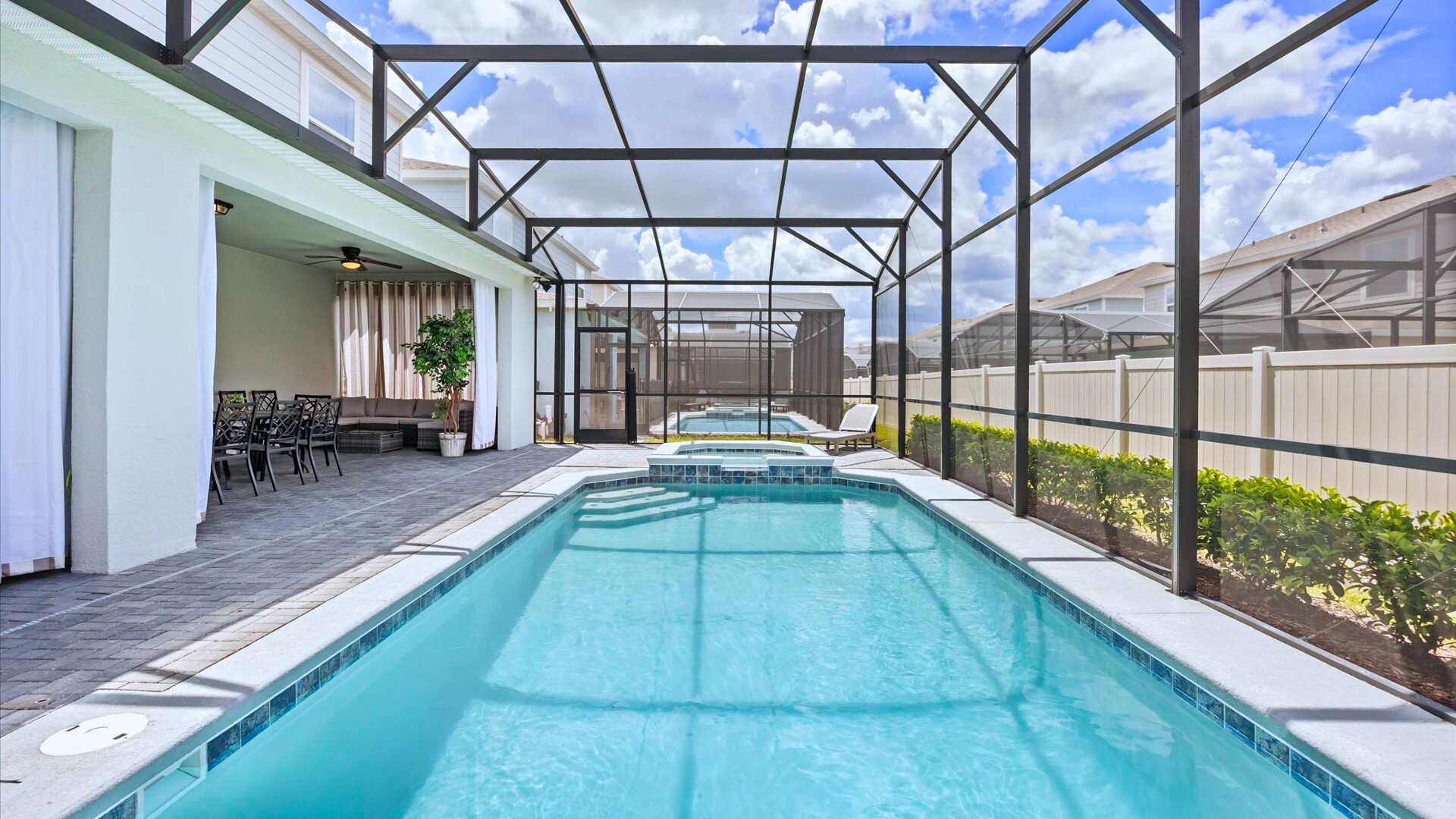 Wide view of the screened-in pool area, showcasing the long swimming pool, hot tub, and the covered patio space with outdoor seating, all under a bright blue sky.
