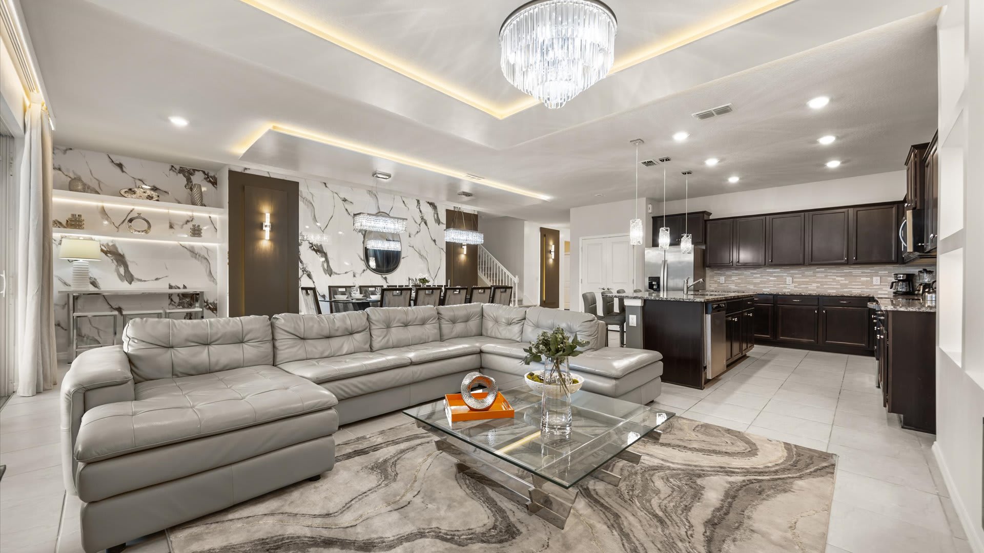 Expansive open-concept living area and kitchen featuring a large grey sectional sofa, a glass coffee table, and a modern chandelier. The kitchen has dark cabinetry and a breakfast bar, all set against white and marble-patterned walls.