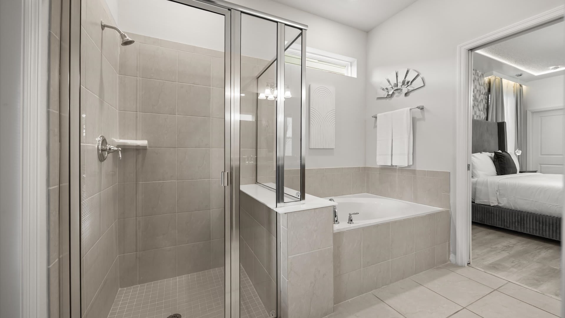 A bright bathroom showcasing a walk-in shower with glass enclosure and light beige tiles, adjacent to a built-in soaking tub, with a view into the master bedroom.
