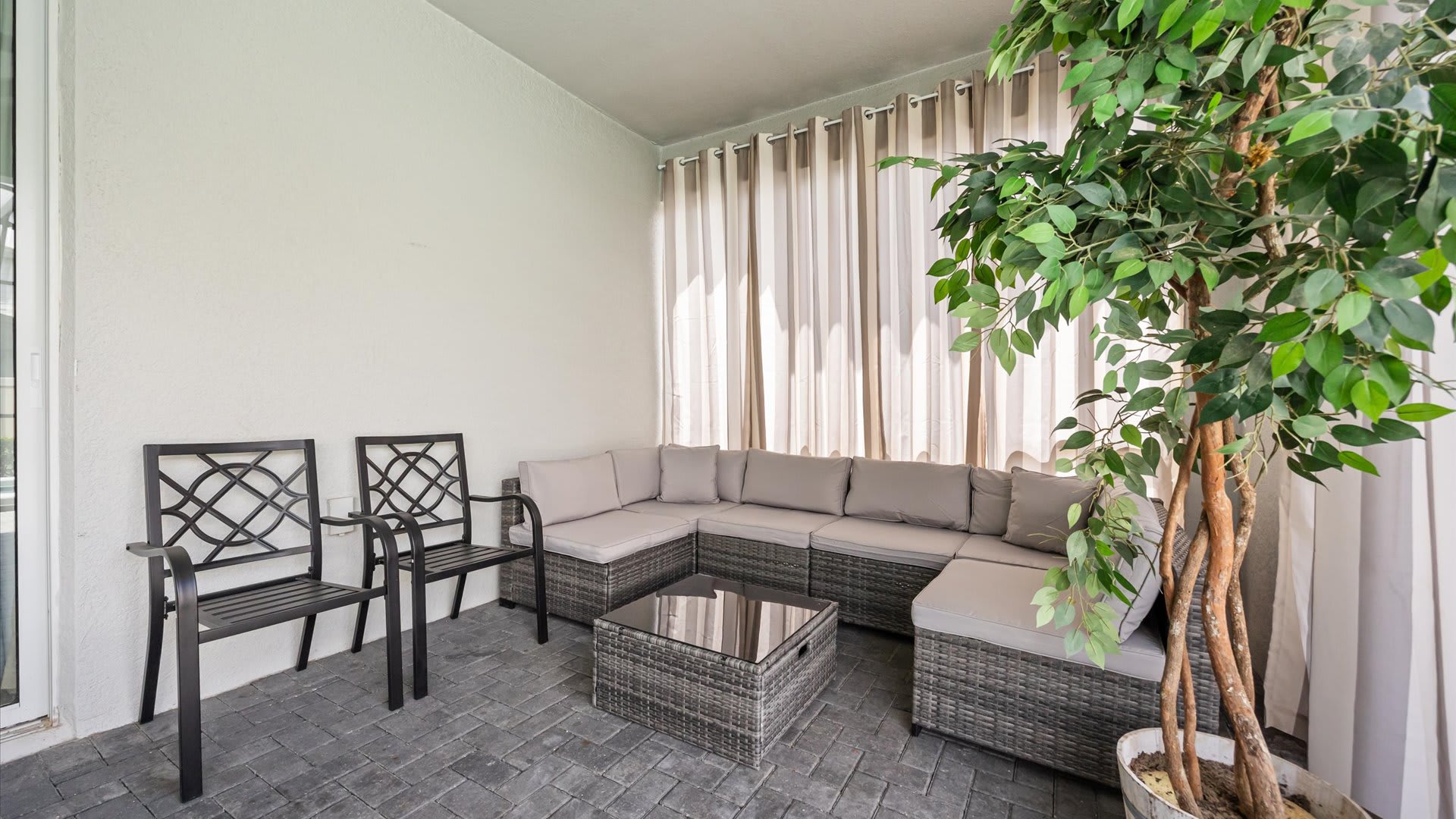 Cozy corner of the covered patio featuring a gray wicker sectional sofa with cushions, two matching chairs, and a glass-top coffee table, surrounded by privacy curtains and a tall artificial plant.