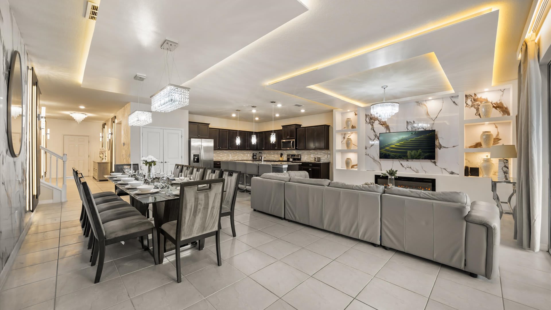 Open-concept living space featuring a long dining table with grey chairs, a grey sectional sofa, and a modern kitchen with dark cabinetry, all under tray ceilings with accent lighting.
