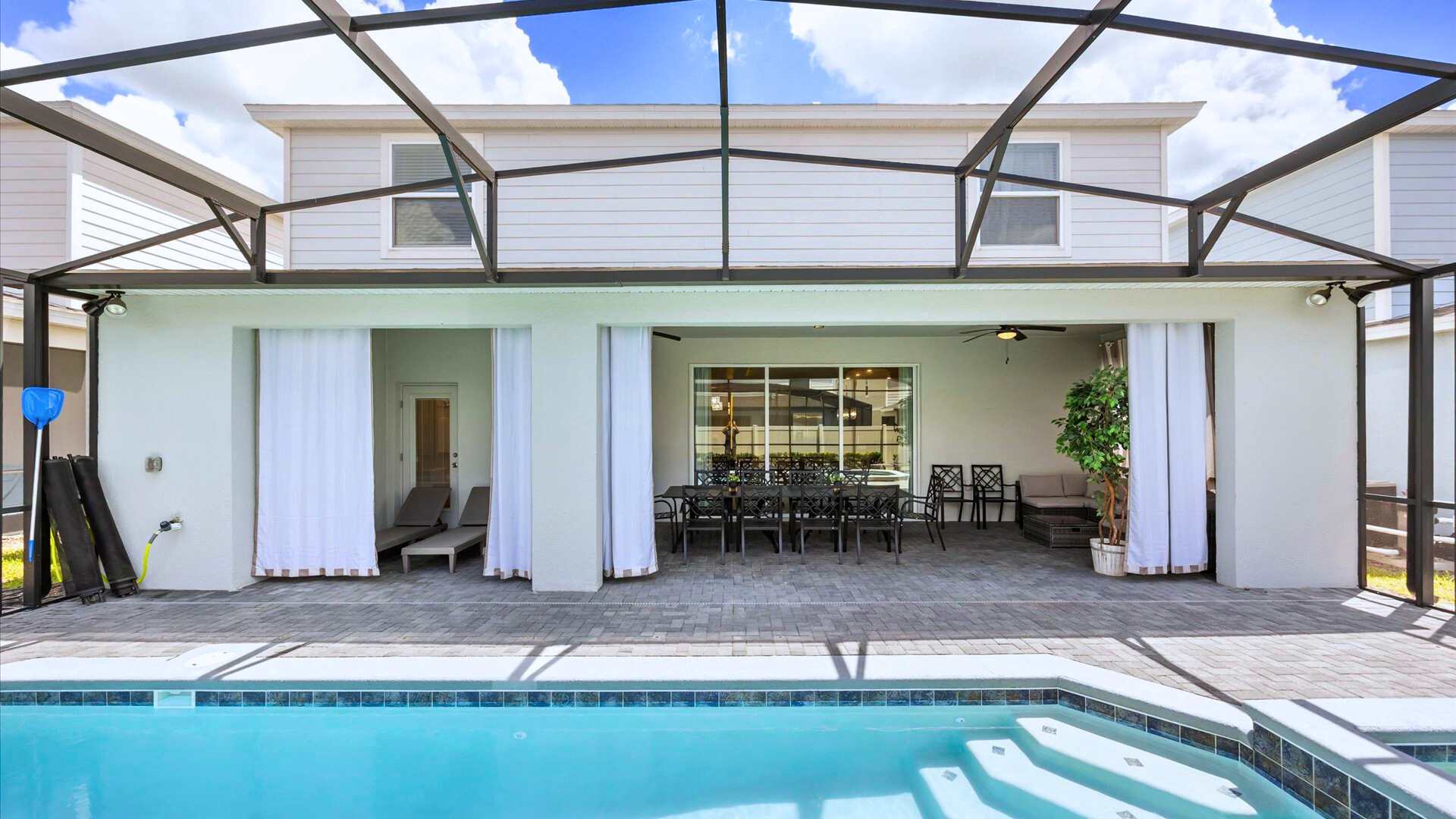 Screened-in outdoor patio area with a large rectangular swimming pool and an adjacent hot tub, surrounded by a paved deck and an outdoor dining table, offering a private oasis.
