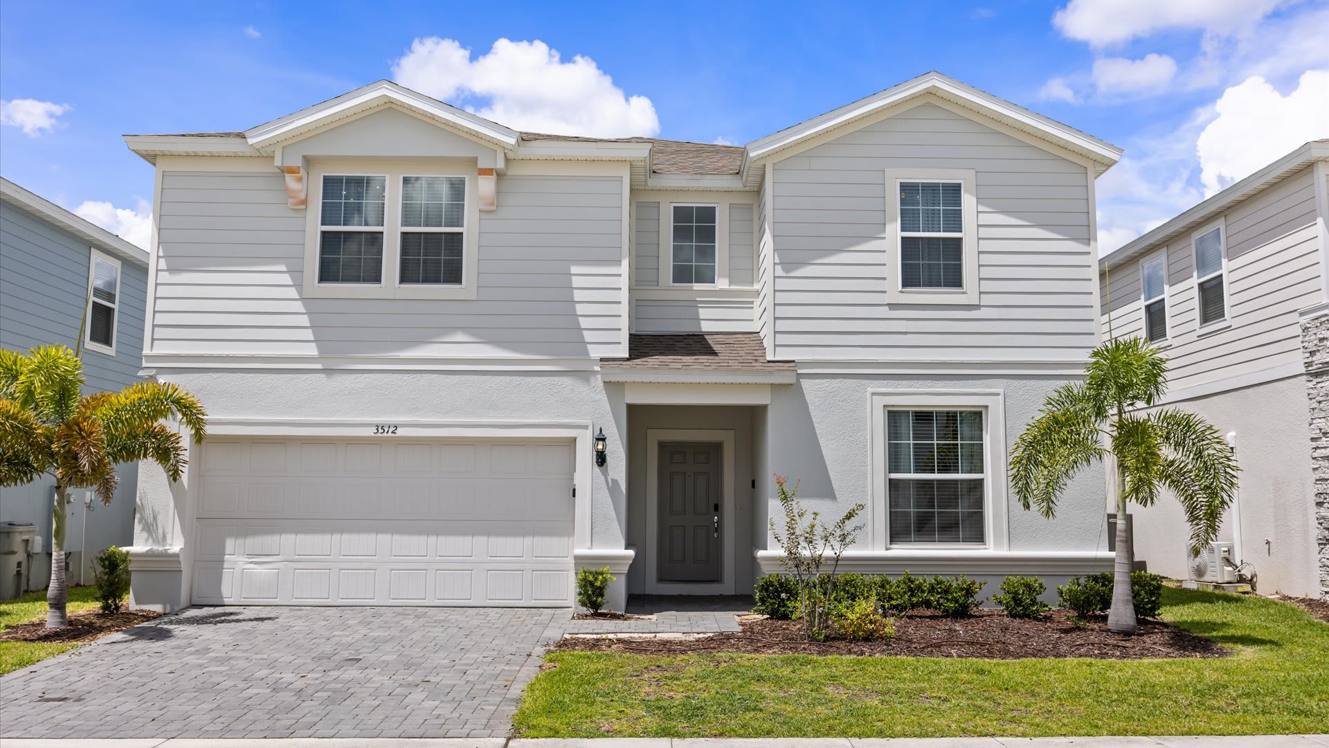 Modern two-story house with light gray siding and white trim, a two-car garage, and a paved driveway, flanked by small palm trees under a blue sky.
