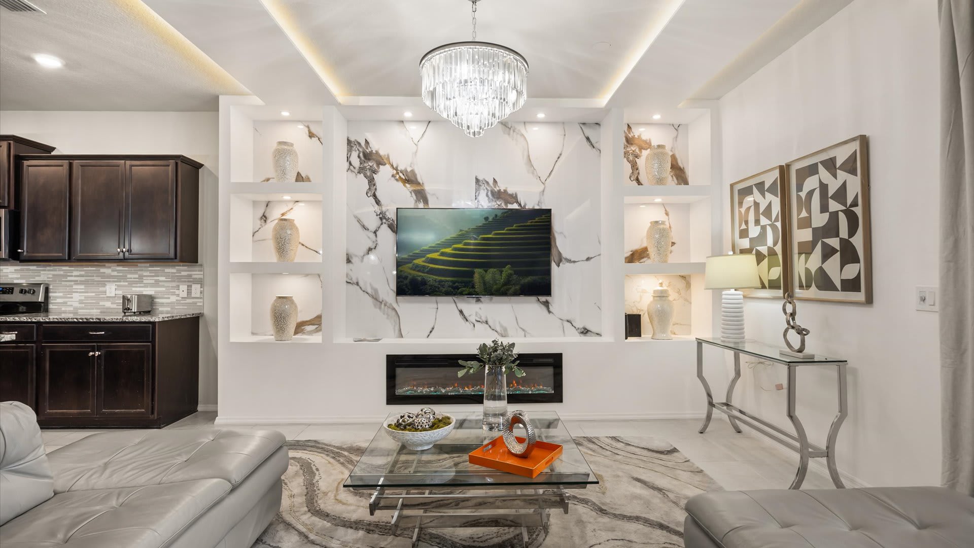 Detailed view of the modern living room featuring a wall-mounted TV set against a white and grey marble-patterned wall with decorative shelves, an electric fireplace, and a glass coffee table.