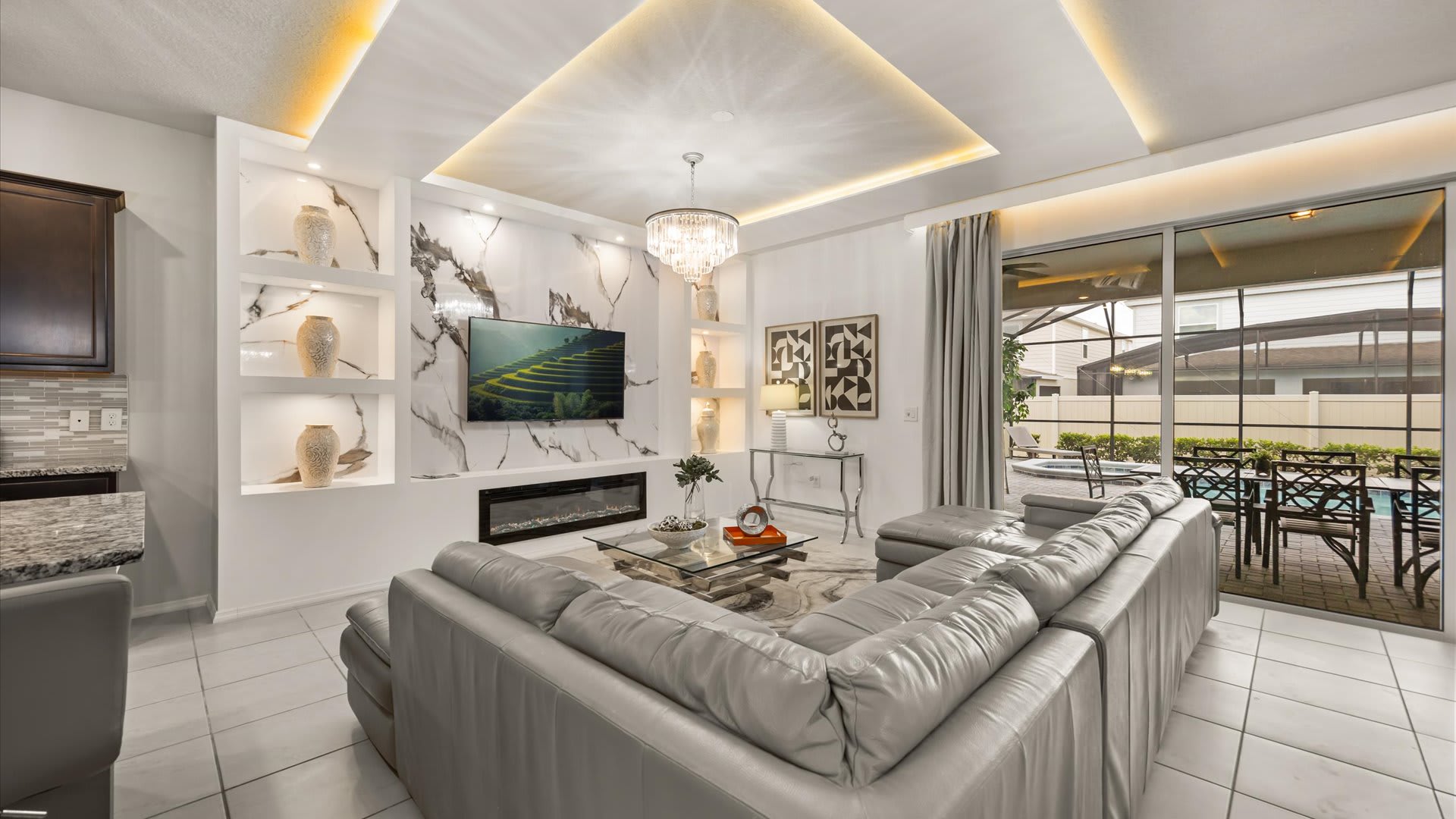 Contemporary living room with a large grey sectional sofa, glass coffee table, and a wall-mounted TV above an electric fireplace, opening to an outdoor patio with a pool.