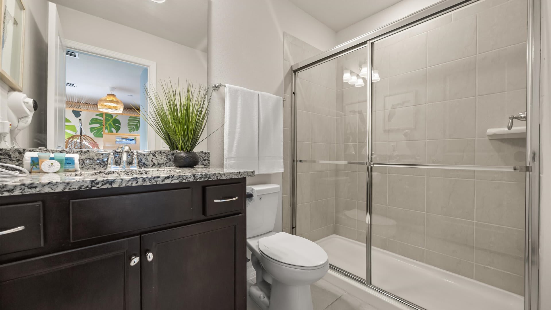 A modern bathroom with a dark wood vanity, speckled granite countertop, white toilet, and a glass-enclosed shower with light beige tiled walls and a plant accent.