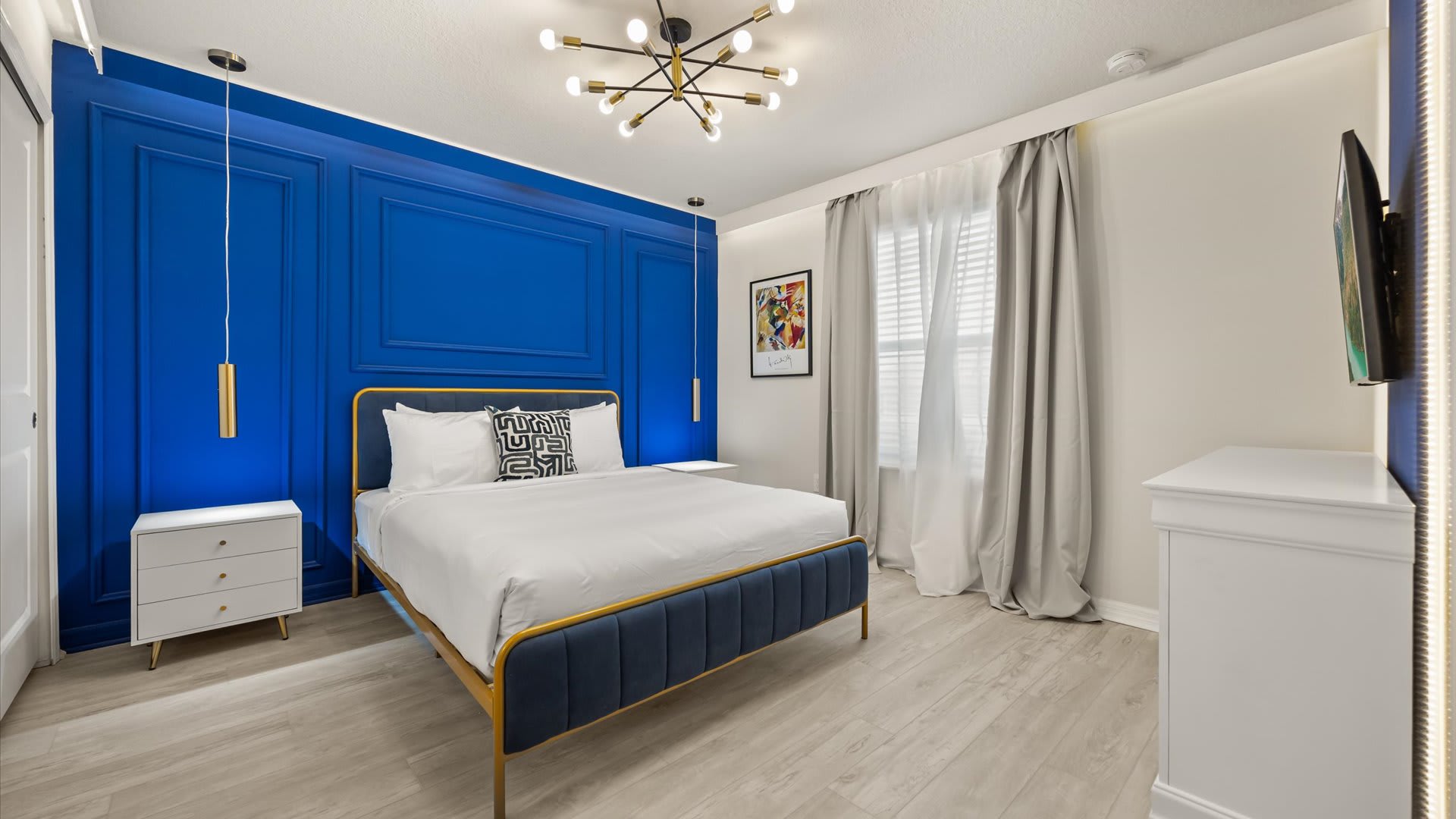 A stylish bedroom featuring a vibrant blue accent wall with decorative paneling, a gold-framed bed with a blue upholstered headboard, and light wood-look flooring, illuminated by a modern sputnik chandelier.