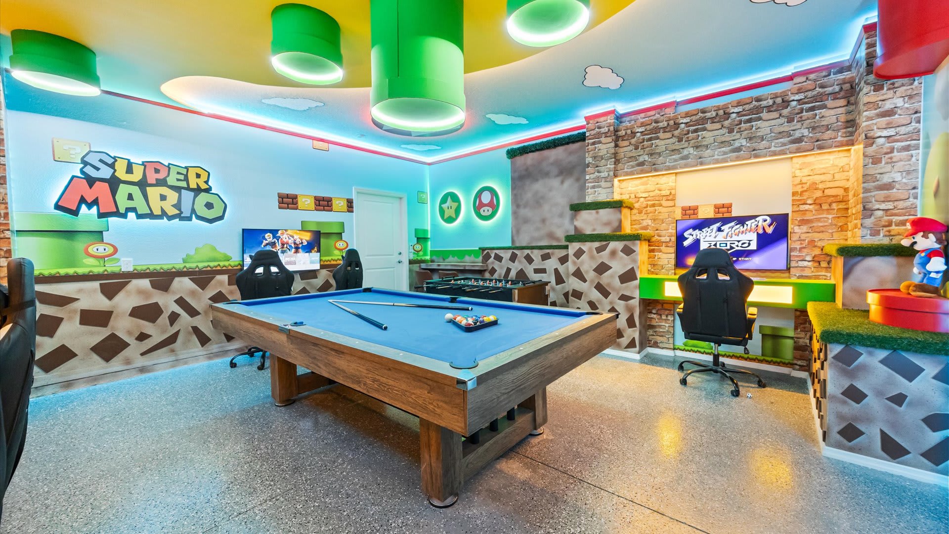 Vibrant Super Mario-themed game room with a blue-felt pool table, two gaming stations with racing chairs, and a foosball table, all under colorful LED lighting and themed decor.