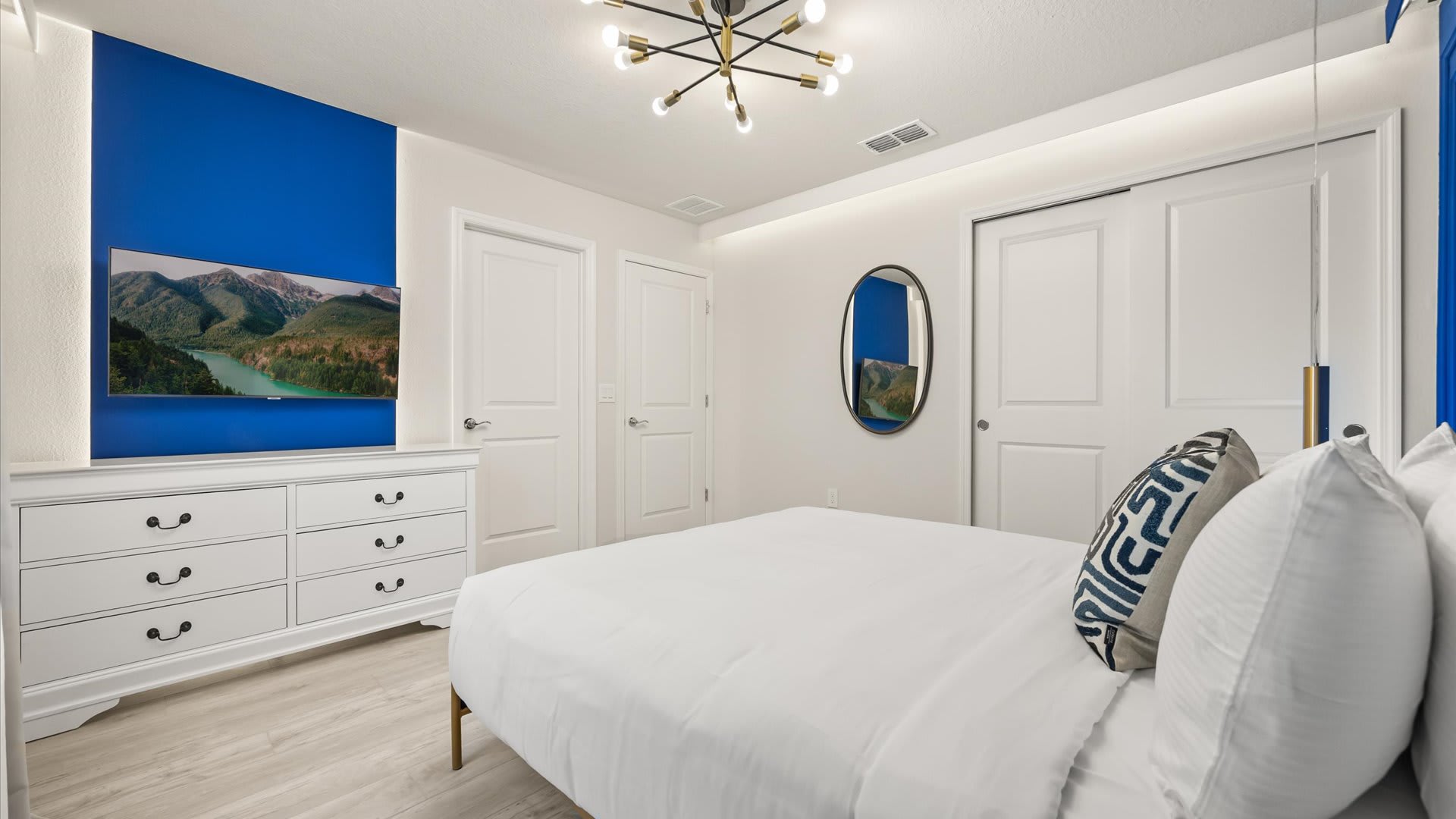 Another perspective of the stylish bedroom, showing a white dresser with a wall-mounted TV, light wood-look flooring, and white double doors, with a glimpse of the blue accent wall.