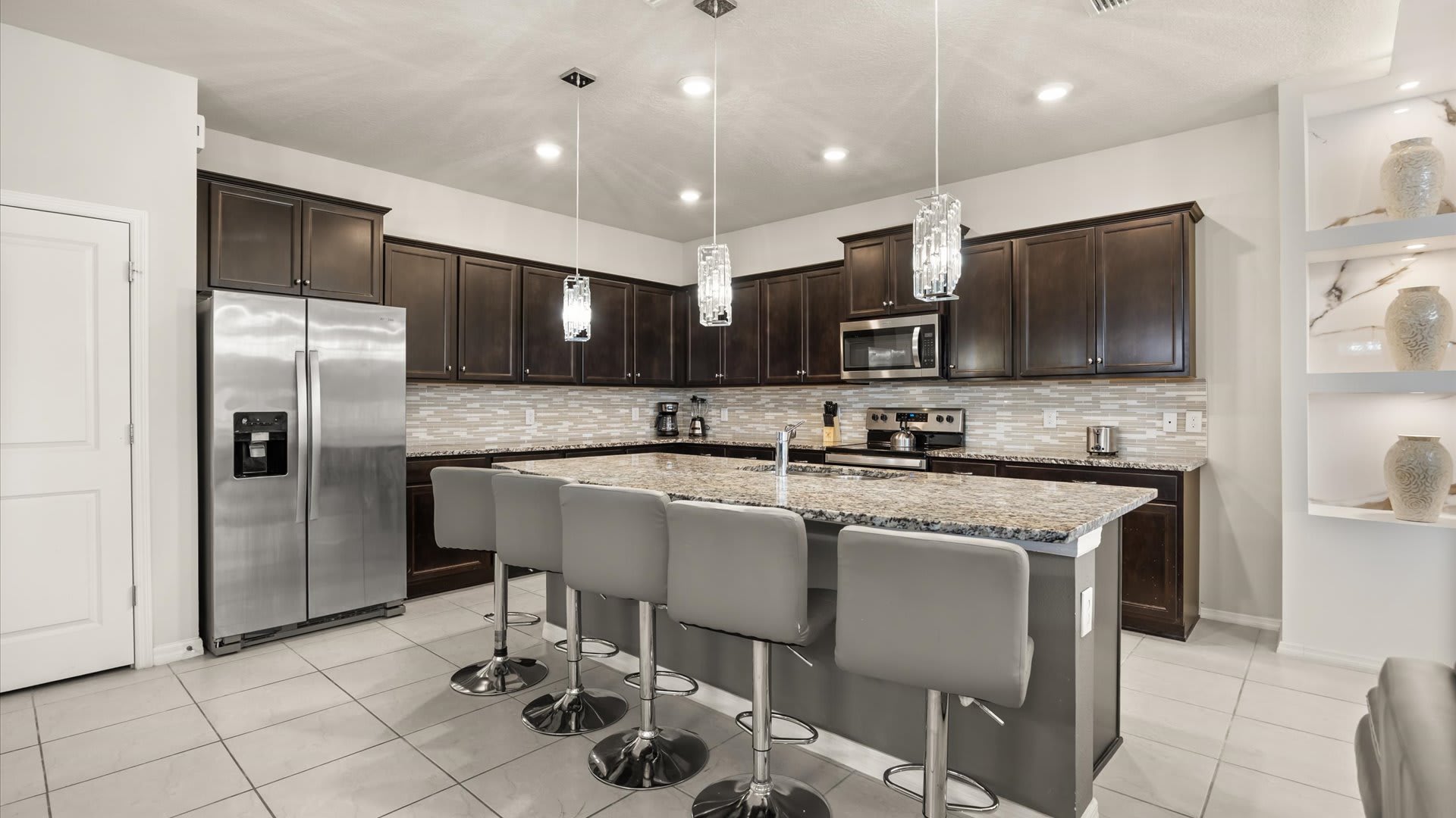 Modern kitchen with dark wood cabinetry, stainless steel appliances, granite countertops, and a large island with four grey bar stools, illuminated by pendant lights.