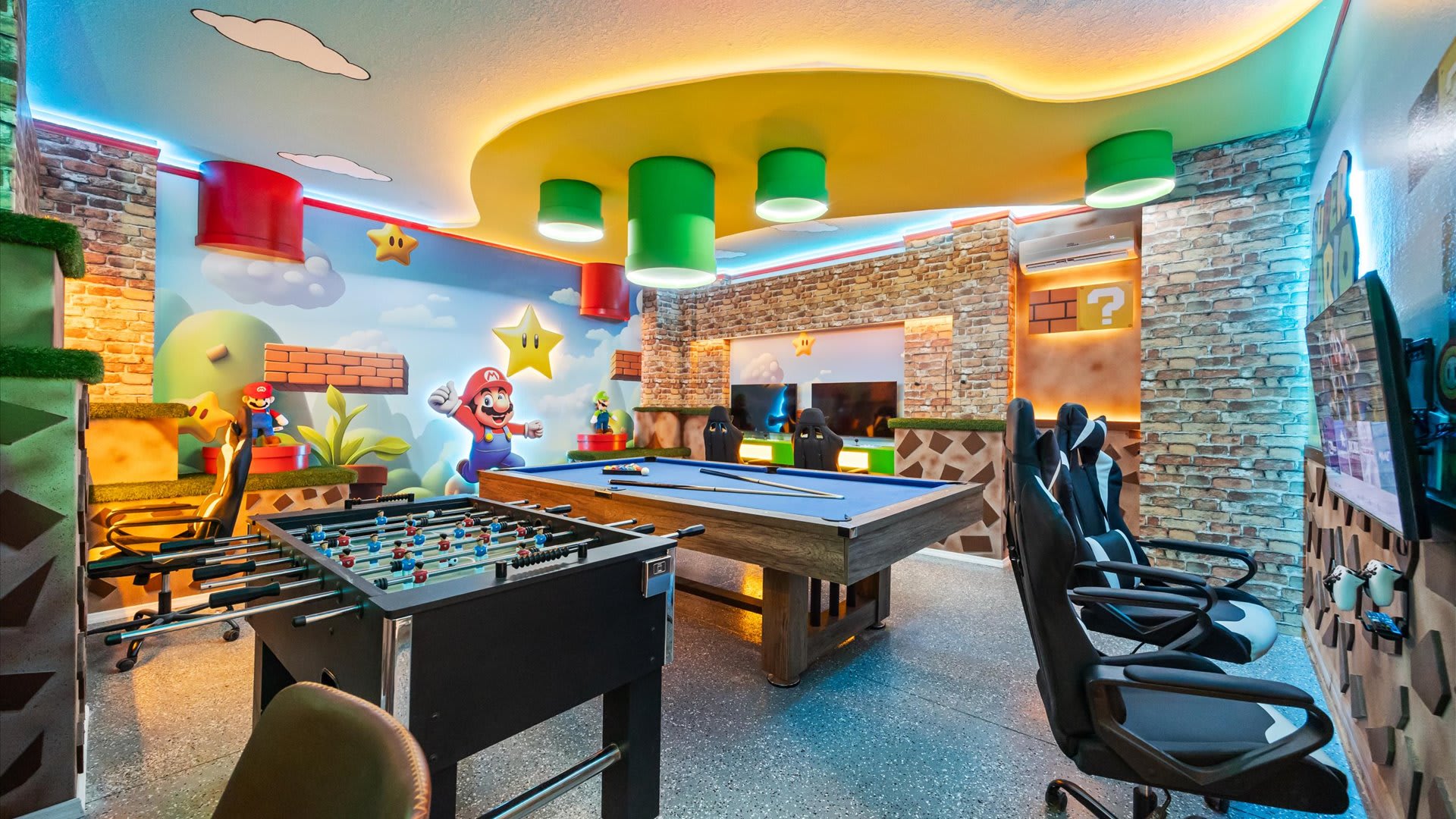 A vibrant Mario-themed game room featuring a pool table, foosball table, and multiple gaming stations with racing chairs. The walls are decorated with brick and character murals, and the ceiling has colorful cove lighting.