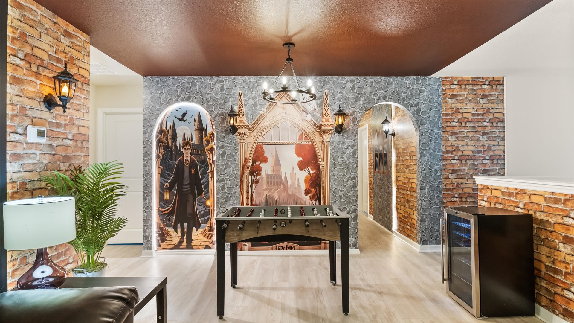 A Harry Potter-themed game room features brick-patterned walls, a dark brown ceiling, and a foosball table at its center. Large arched murals depict Hogwarts scenes, illuminated by wall sconces and a central chandelier.