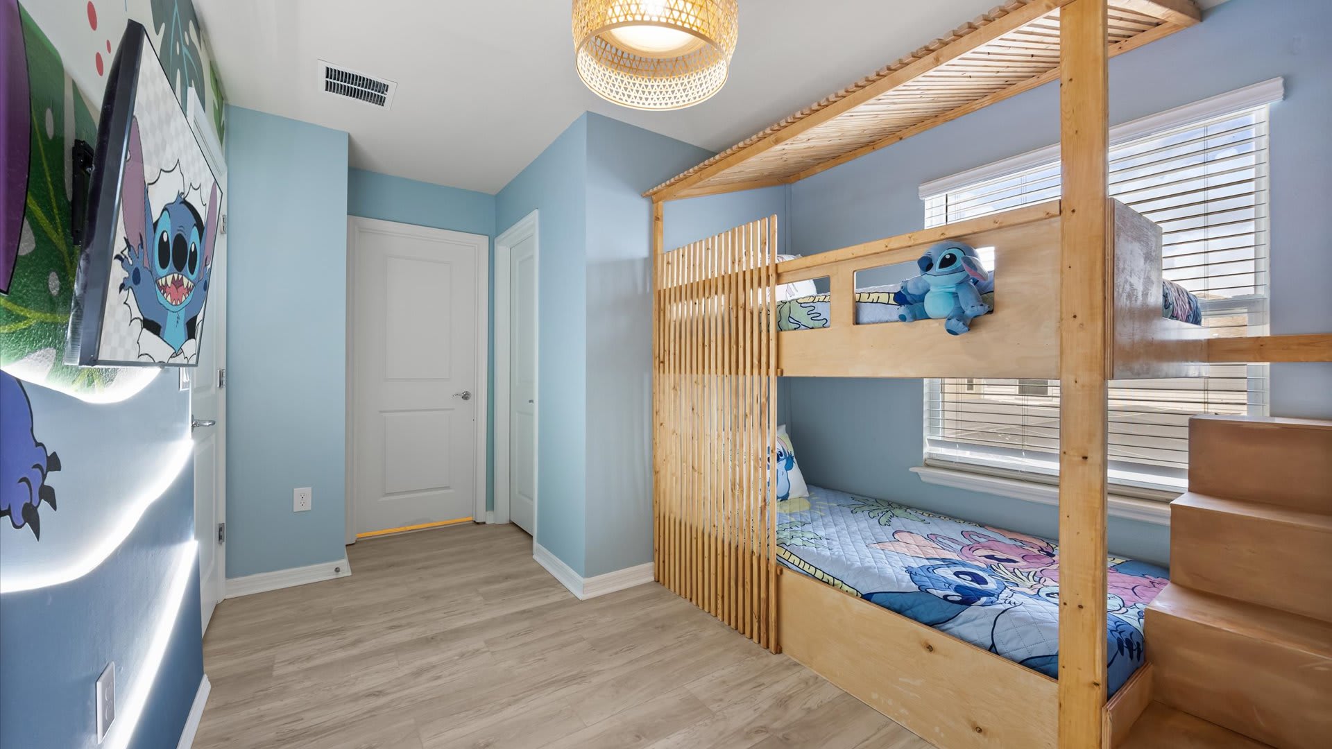 A vibrant children's bedroom featuring a light wood bunk bed with a Lilo & Stitch theme, light blue walls, and a wall-mounted TV displaying character art.