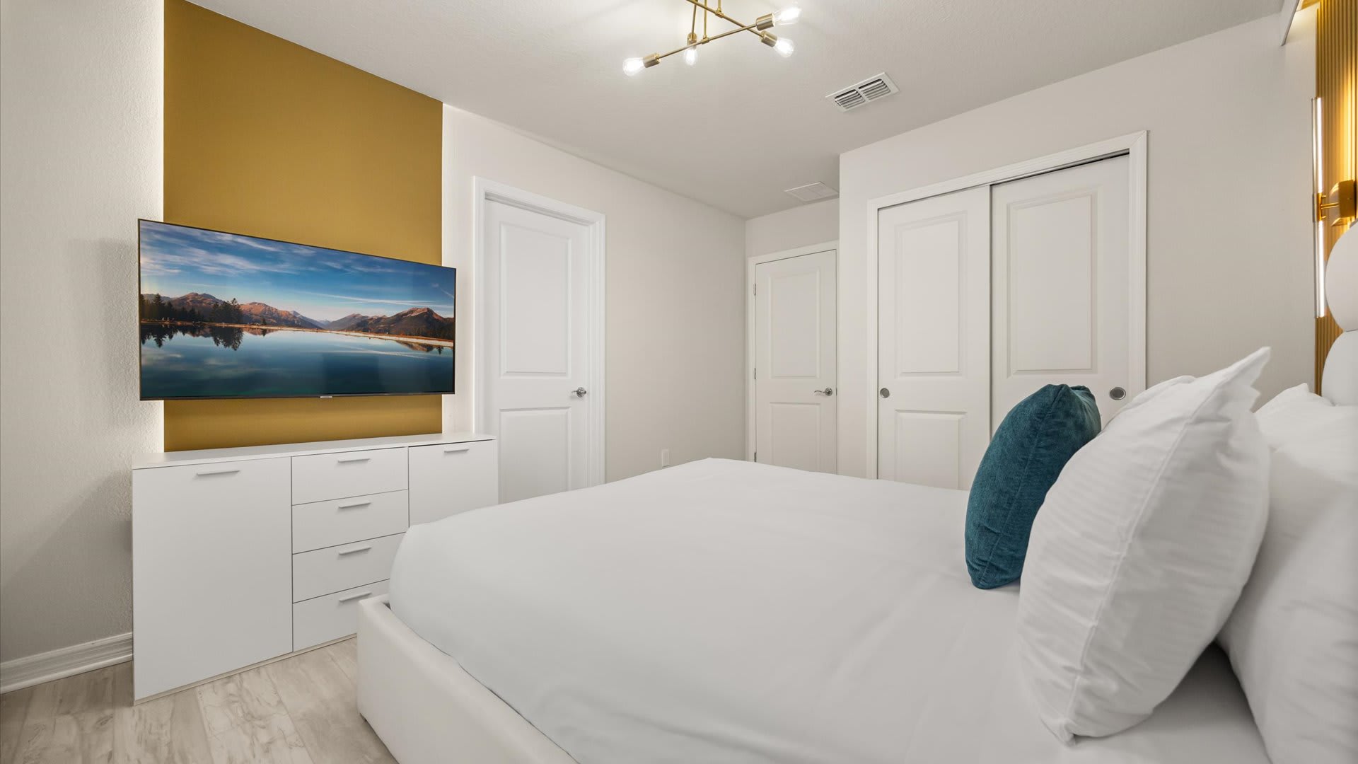 Another perspective of the bright bedroom, showcasing a white dresser with a wall-mounted TV displaying a lake scene, and a closet with white sliding doors.