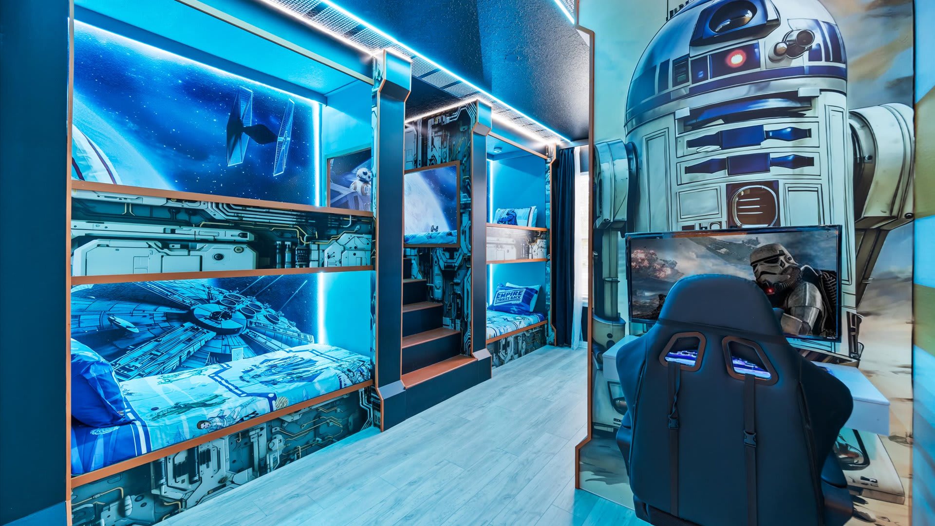 Immersive Star Wars-themed bunk room with multiple custom-built bunk beds, each featuring unique space-themed murals and integrated blue LED lighting. A gaming station with a monitor and chair is visible.