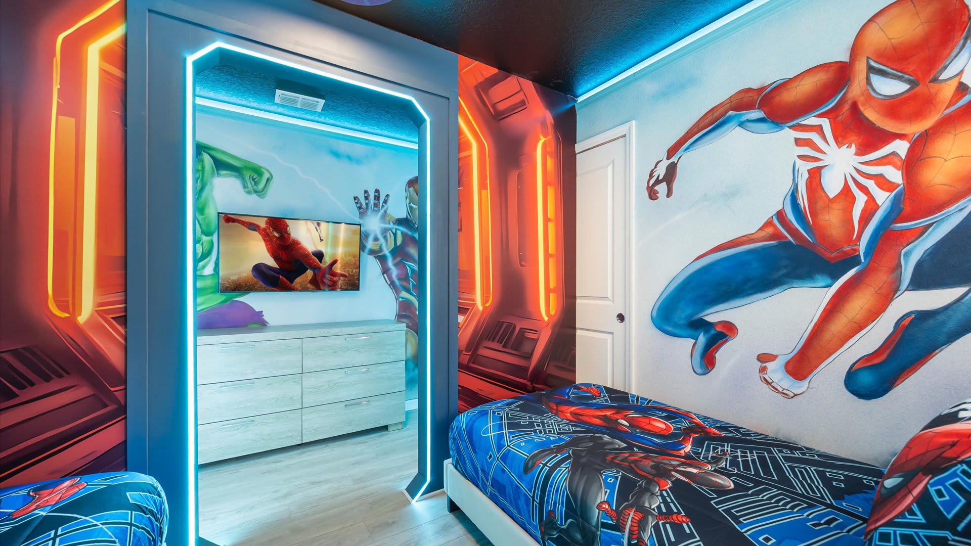 A vibrant Marvel-themed bedroom featuring a large Spider-Man mural on the wall, a bed with themed bedding, and a doorway with glowing blue LED lights leading to another area with a TV and dresser.