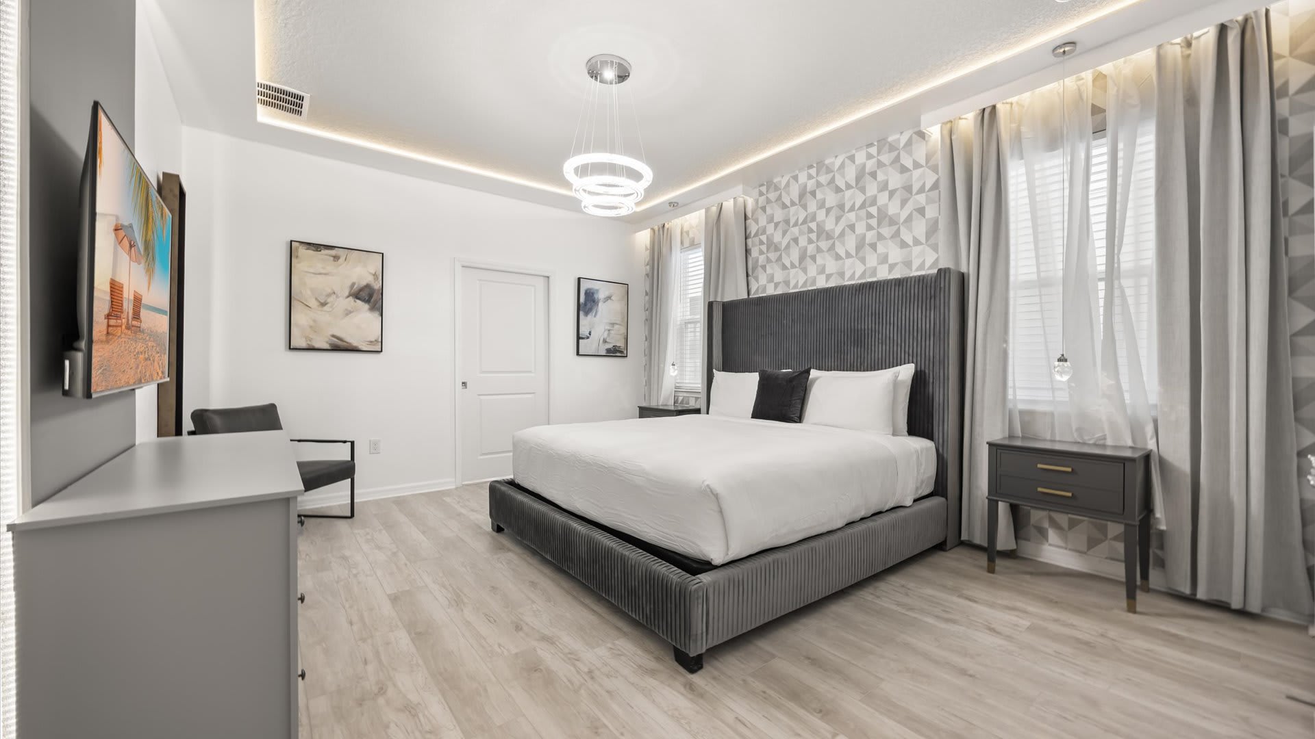A modern master bedroom with a large gray upholstered king-size bed, light wood-look flooring, and a geometric patterned accent wall, illuminated by a contemporary chandelier and recessed lighting.