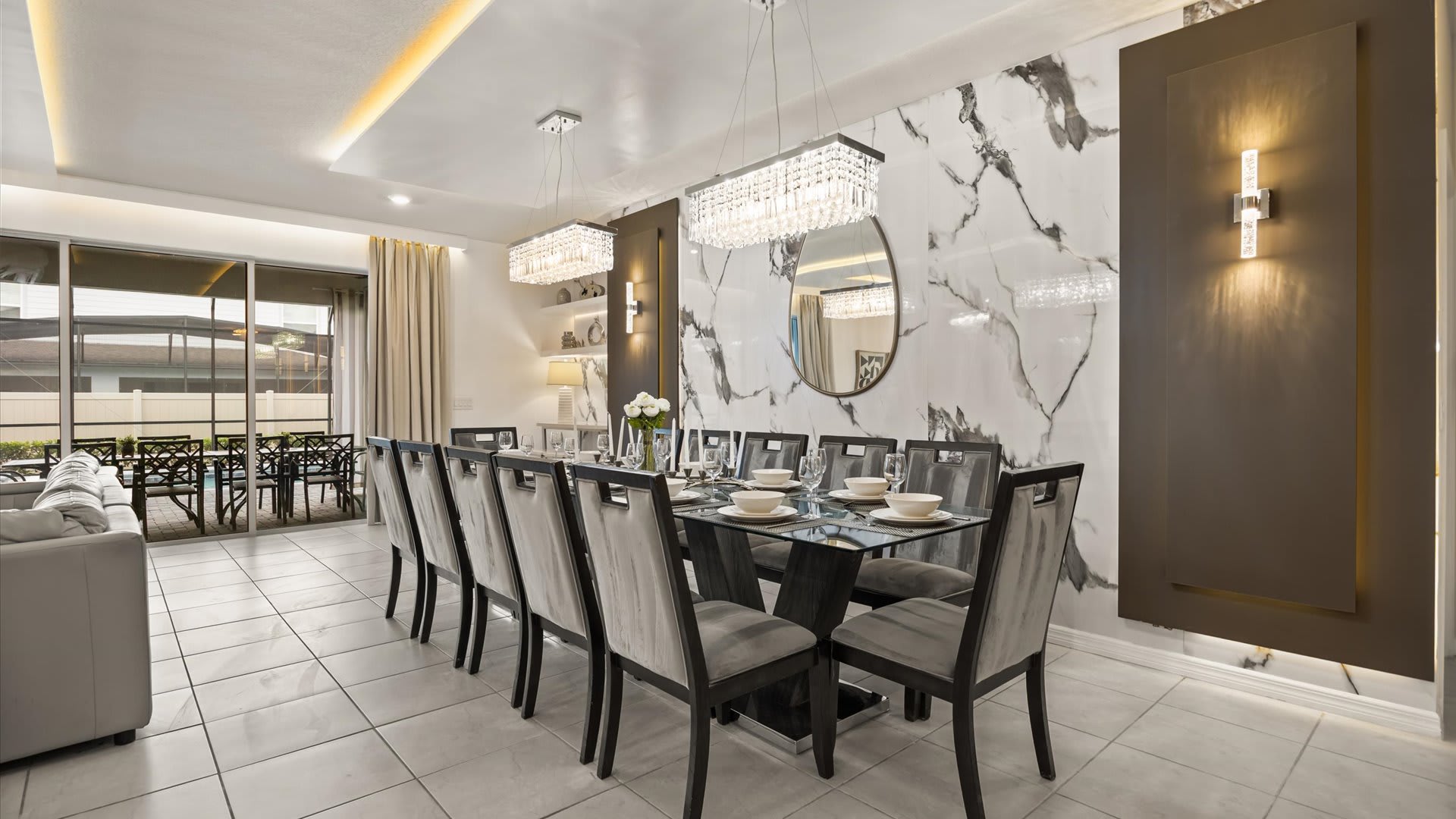 Elegant dining area featuring a long glass-top table with numerous grey upholstered chairs, illuminated by two modern crystal chandeliers, set against a striking marble-patterned accent wall.