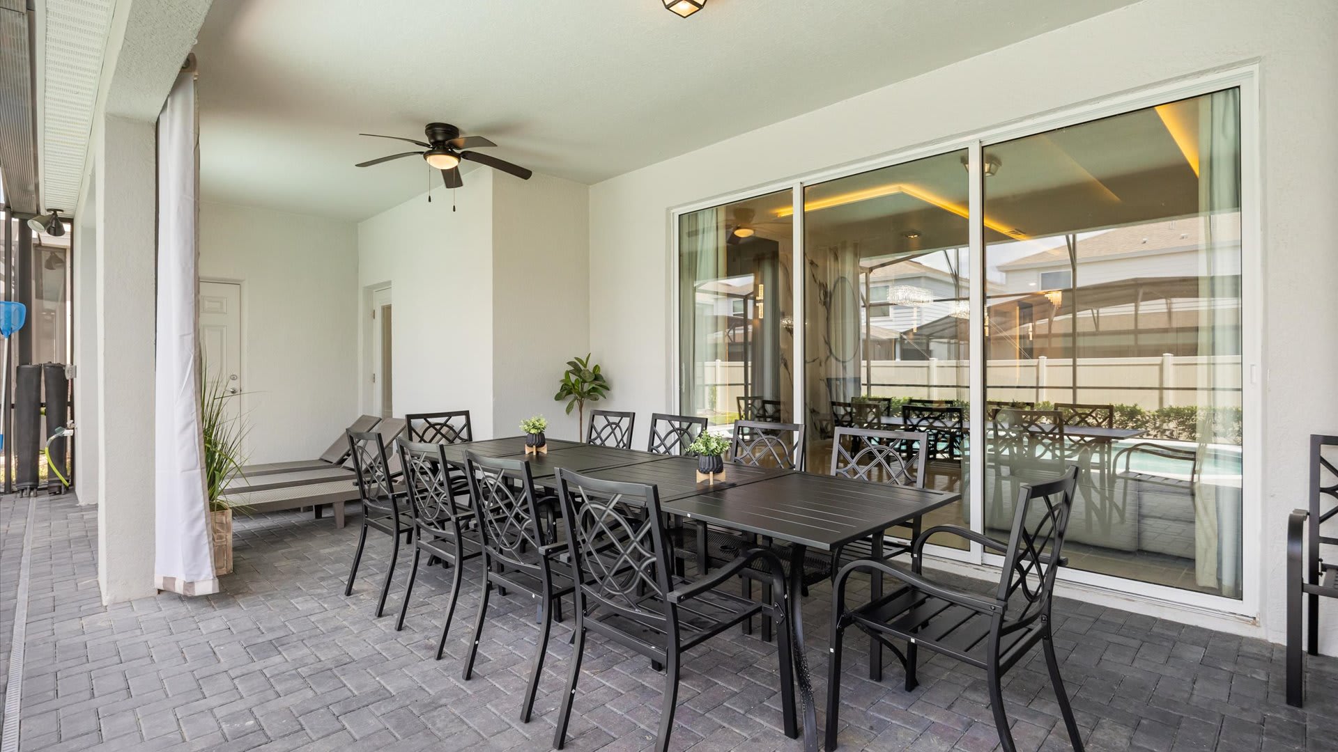 Covered outdoor patio area with a large black metal dining table and chairs, a ceiling fan, and sliding glass doors leading into the house, overlooking the pool area.