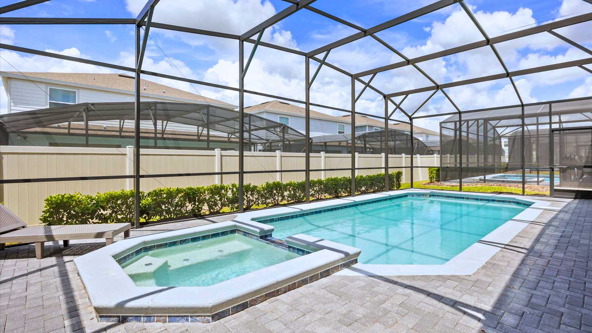 Screened-in outdoor pool area featuring a rectangular swimming pool and an adjacent hot tub, surrounded by paver decking. A lounge chair is visible, and a low hedge borders the fence in the background.