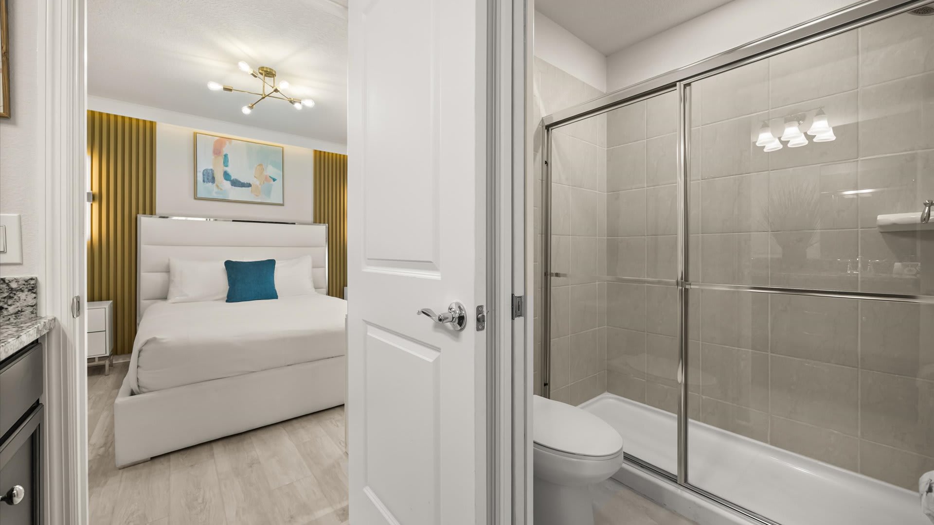 A partial view of a modern bathroom, showing a white toilet and a glass-enclosed shower with light tiled walls, visible from an adjacent bedroom with a white bed.