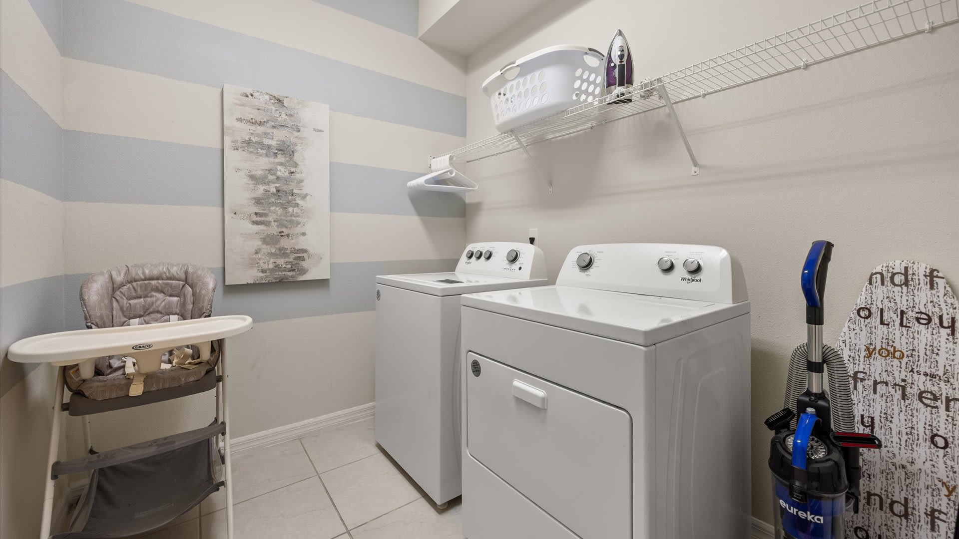 A functional laundry room features a white top-load washer and front-load dryer, with a wire shelf above holding laundry supplies. The walls are painted with light blue and white horizontal stripes, and a high chair is visible on the left.