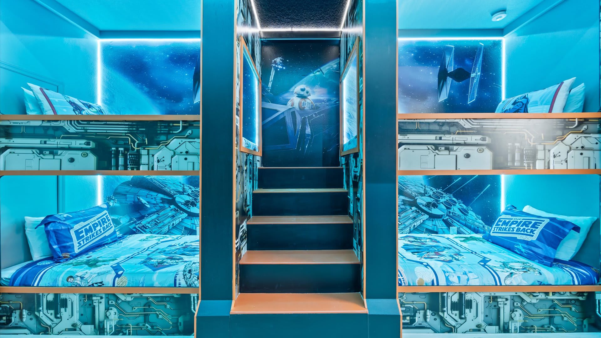 A Star Wars-themed bunk room with multiple bunk beds featuring blue LED lighting, themed bedding, and murals of spaceships and characters on the walls, centered around a short staircase.
